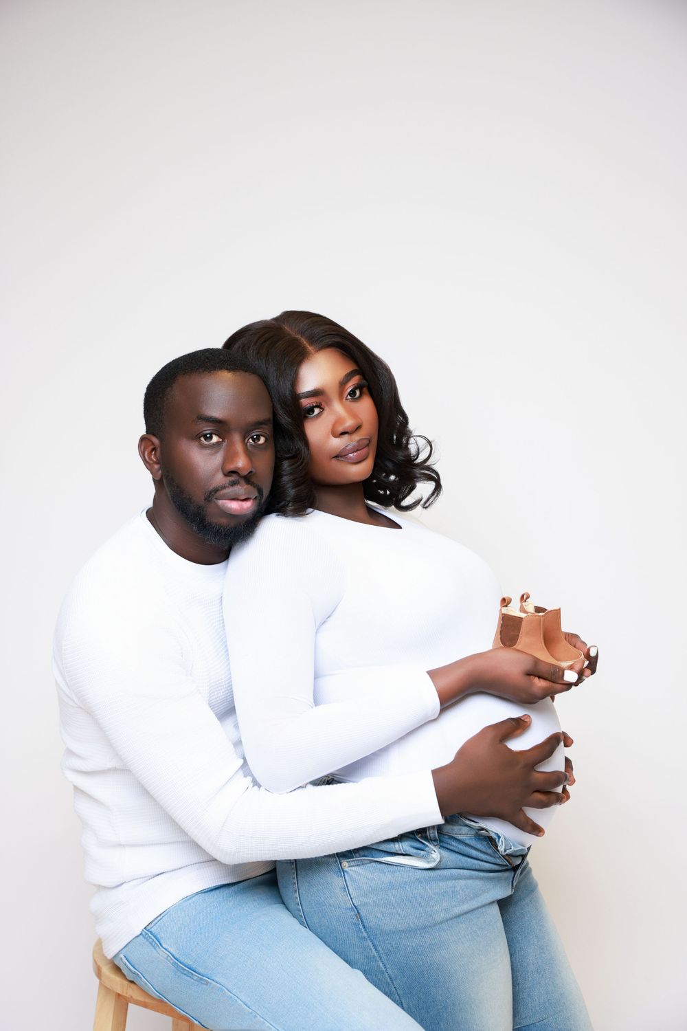 An intimate maternity portrait of a couple in white tops and jeans posed against a white background.