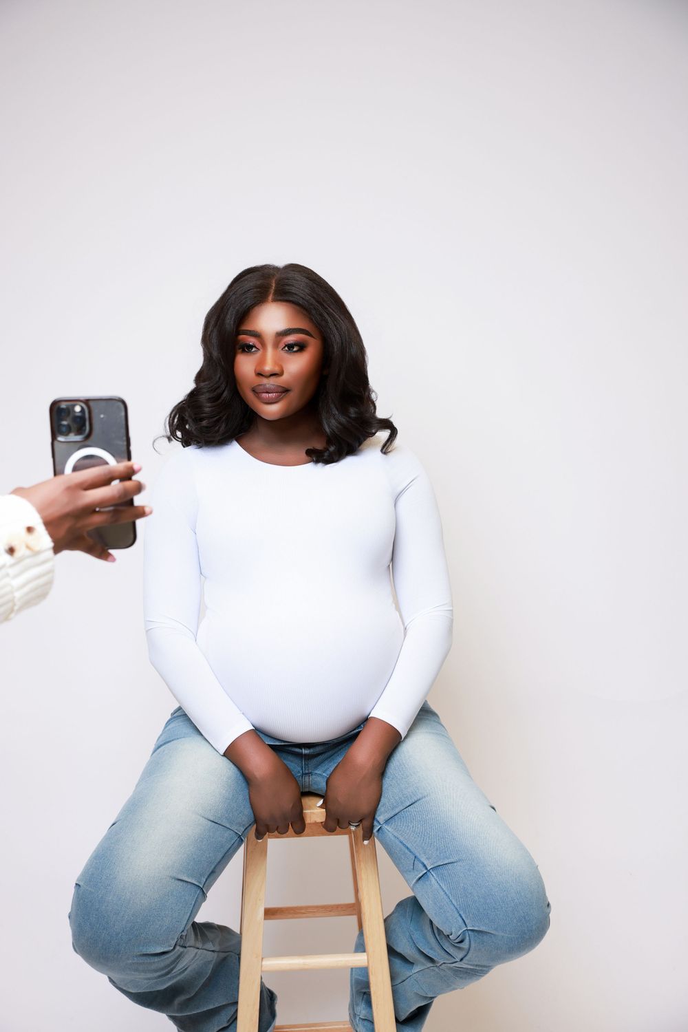 Expectant mother in white sweater and jeans poses on wooden stool while someone takes her photo.