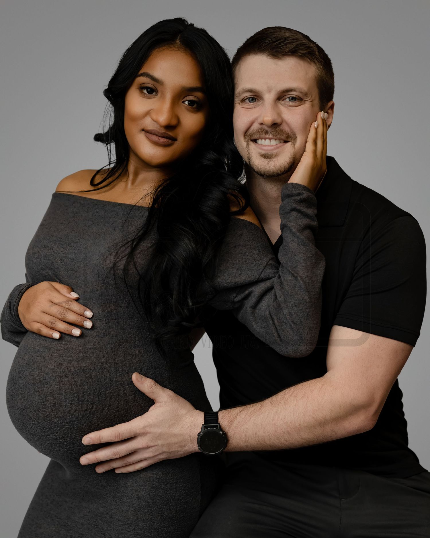 Expecting couple poses together for a maternity photo wearing black outfits against a gray background.