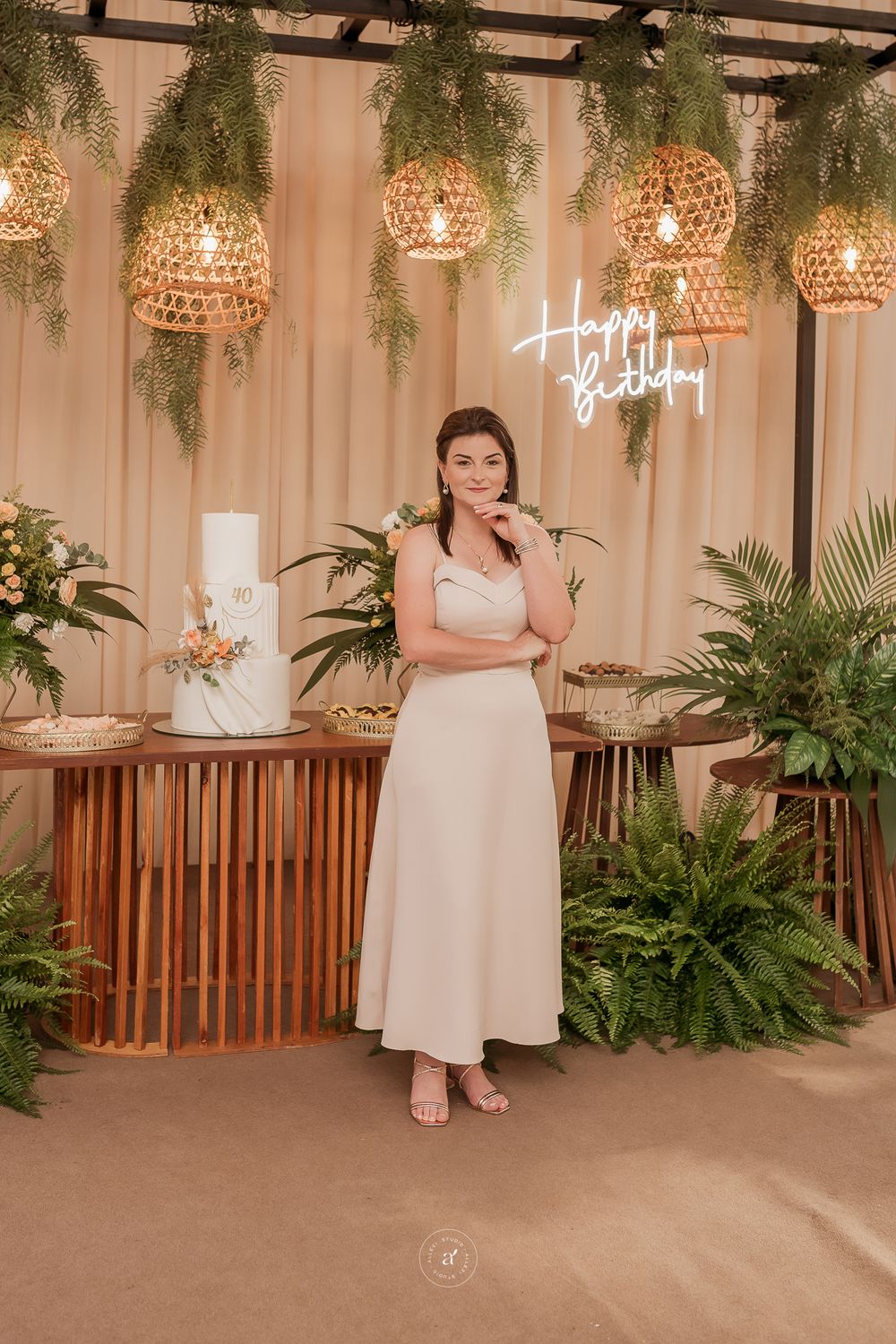 A wedding reception setup with hanging rattan pendant lights, tropical greenery, and a white tiered cake on a wooden display.