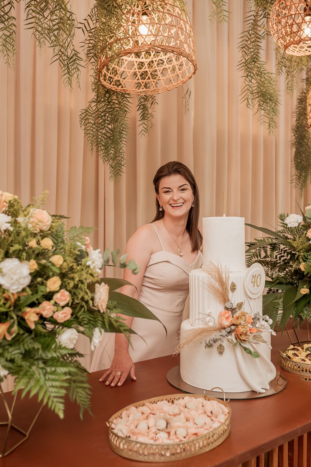 Elegant wedding celebration setup featuring a white tiered cake, woven pendant lights, and soft peach floral arrangements.