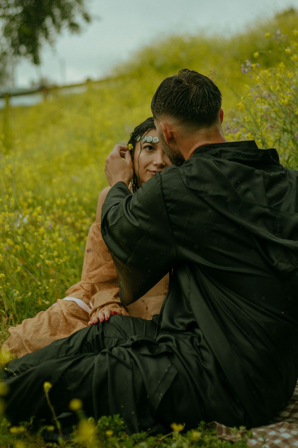 Husband romantically putting a flower in wife's hair while they sit in the rain in a field of flowers in Los Angeles, California.