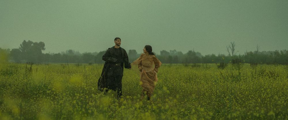 Romantic couple walk together through a misty green landscape with tall grass and a moody atmospheric sky.