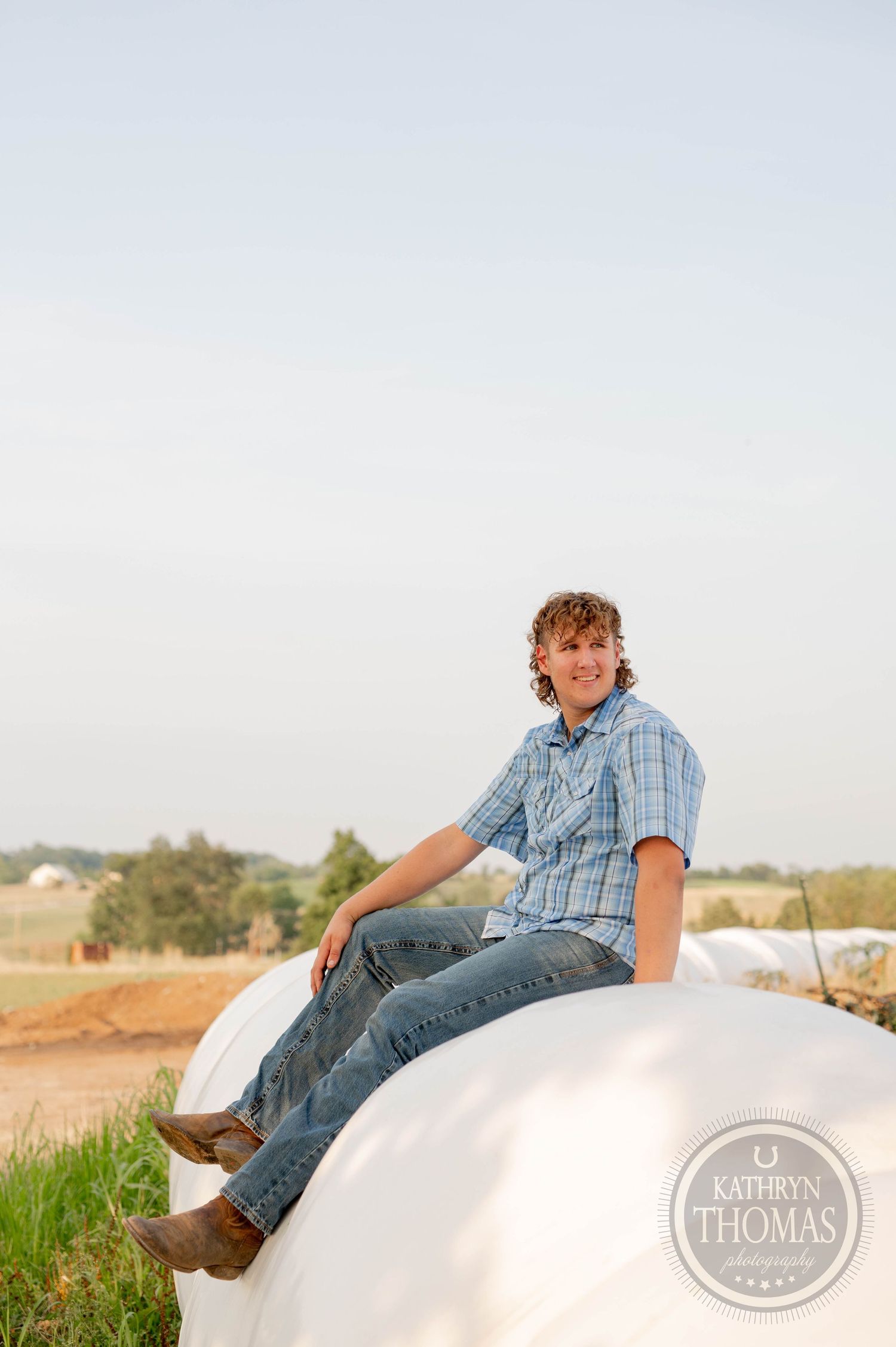 A person sitting on a large white round hay bale in a rural field at dusk.