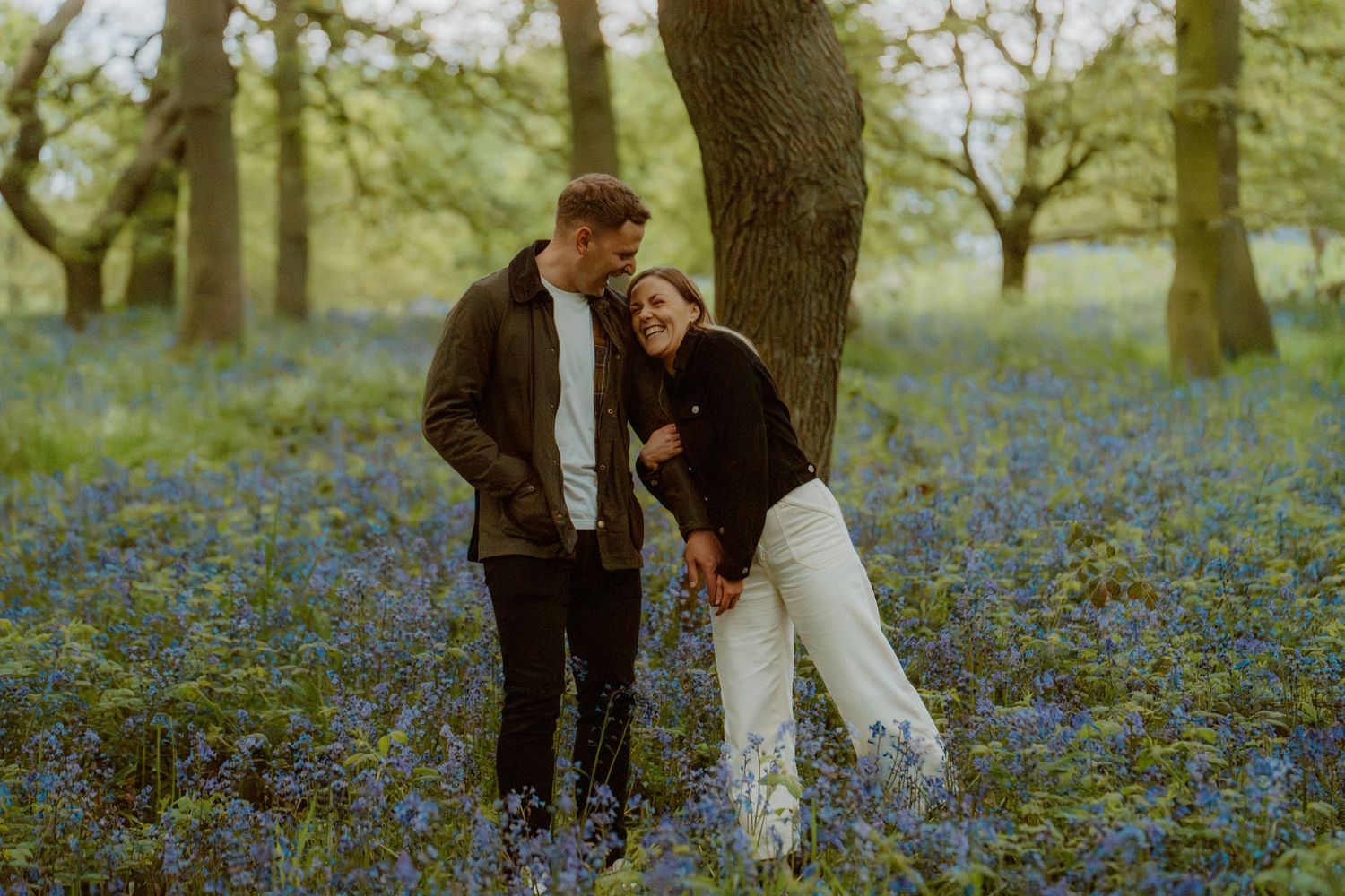 A couple stands together in a dreamy forest of blooming bluebells during golden hour.