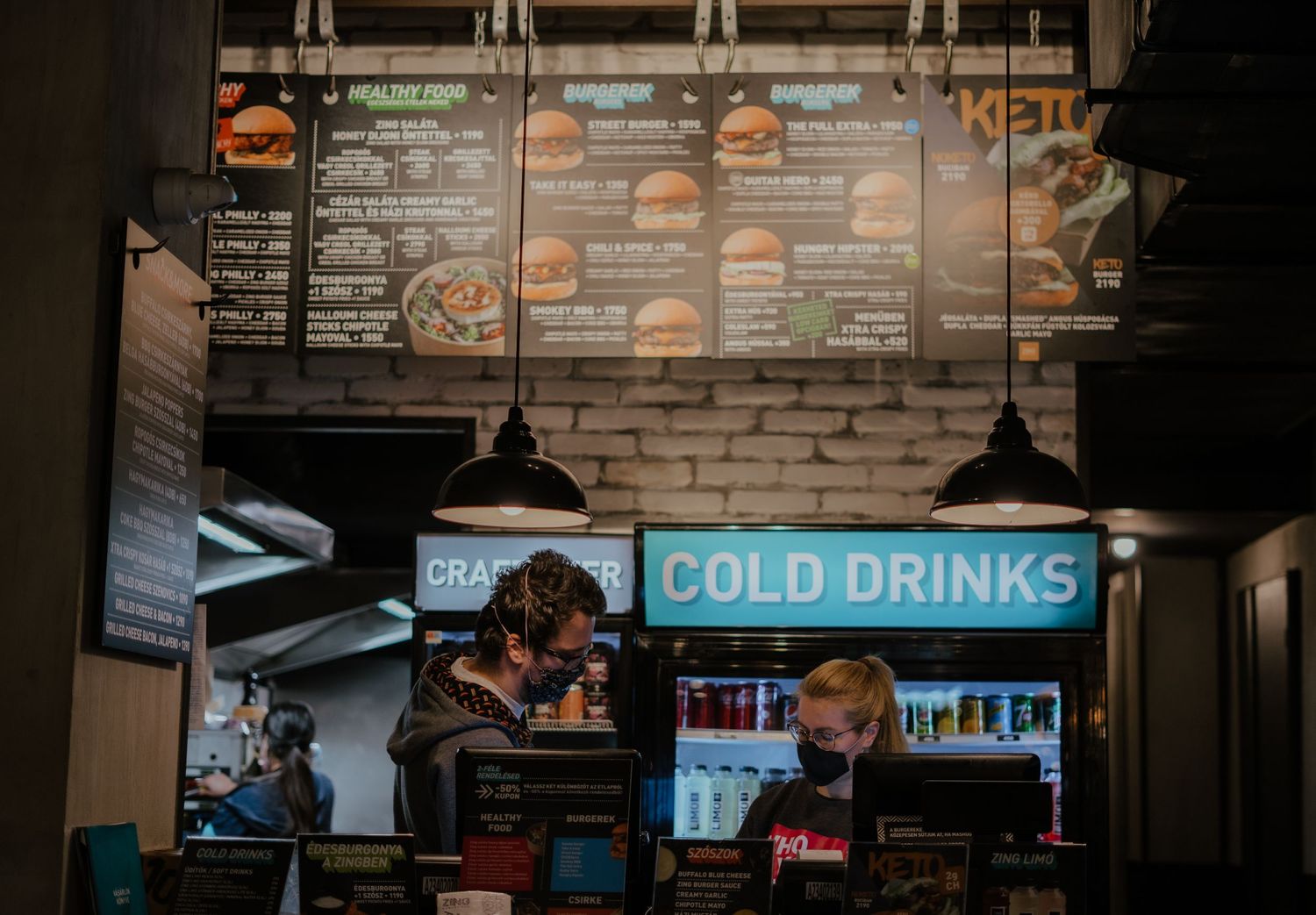Dark cafe interior with illuminated menu boards and 'COLD DRINKS' neon sign hanging above service counter.