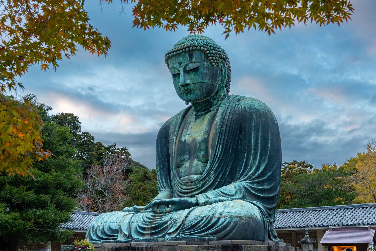 The Great Buddha of Kamakura sits peacefully under autumn leaves and cloudy skies in Japan.