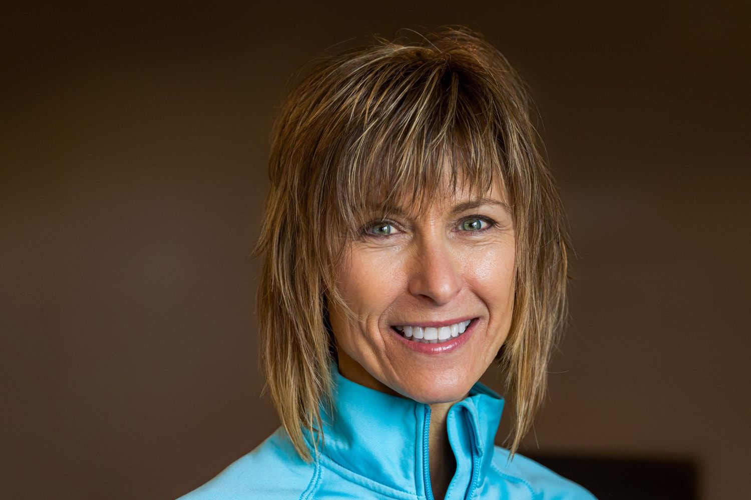 Portrait of someone wearing a bright turquoise athletic jacket, with a warm smile against a dark brown background.