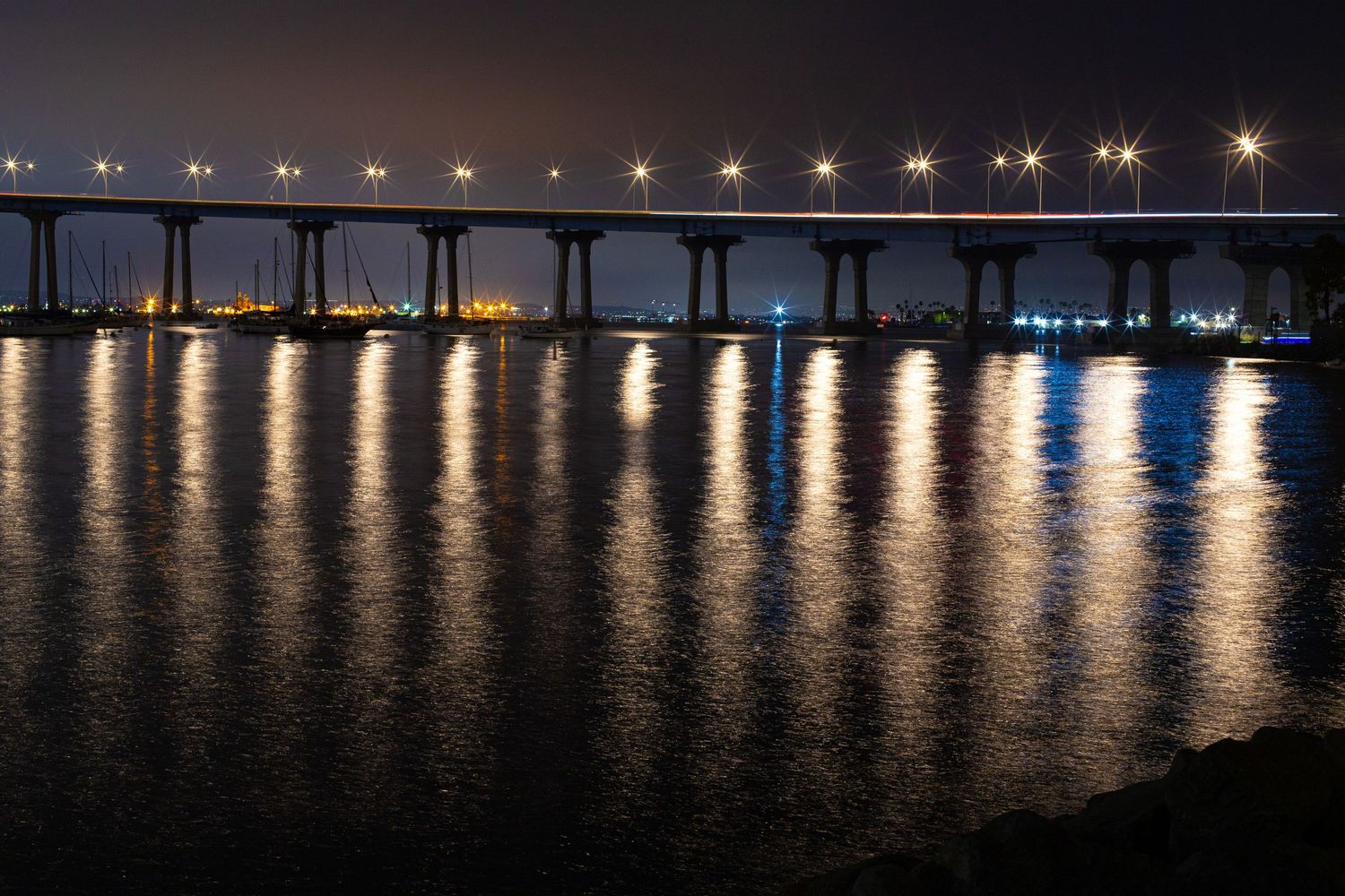 Illuminated Coronado bridge spans across dark water at night with lights reflecting on the surface.