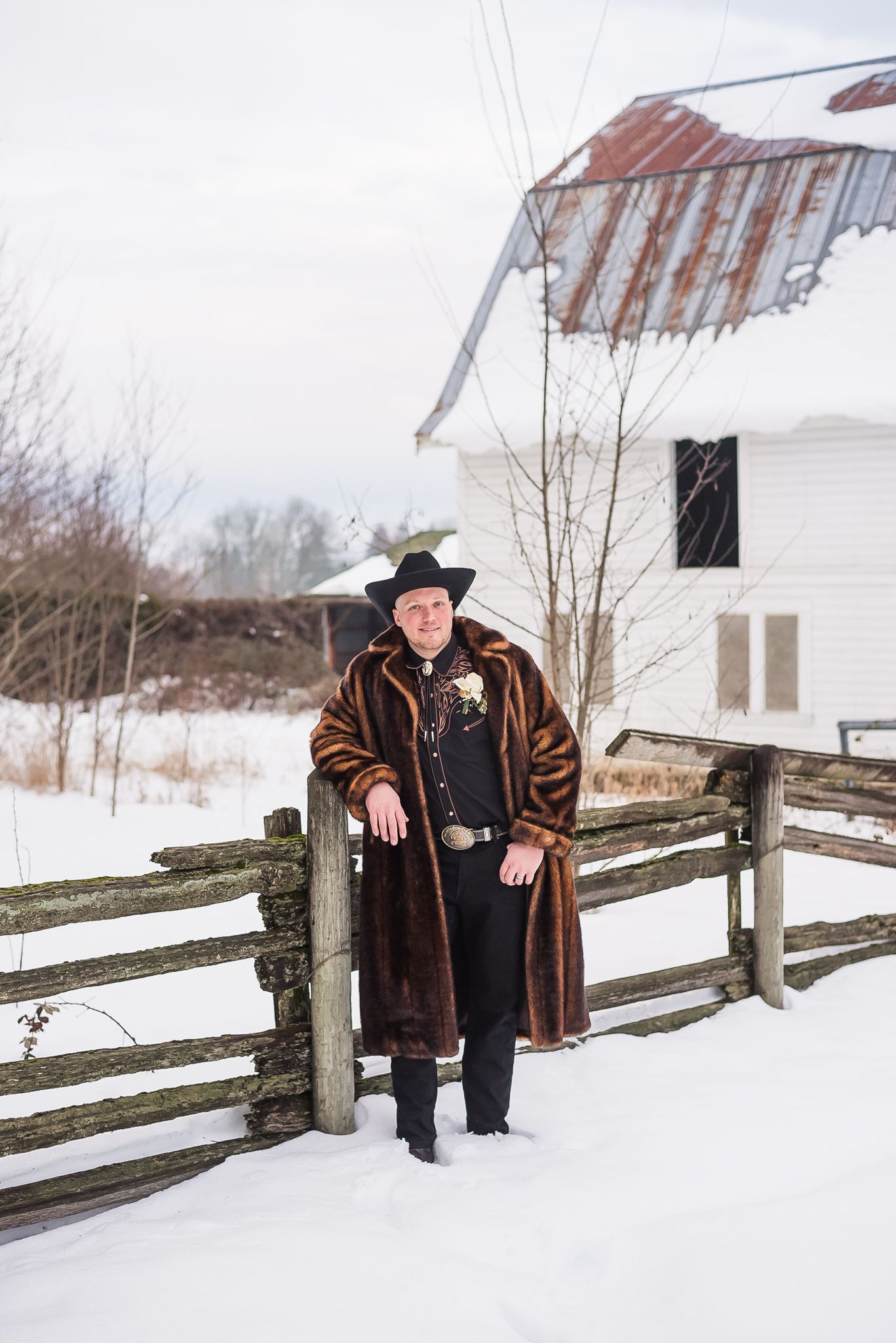 A man, wearing a cowboy hat and faux fur jacket, poses by a fence at his rustic barn wedding venue in Langley, BC.