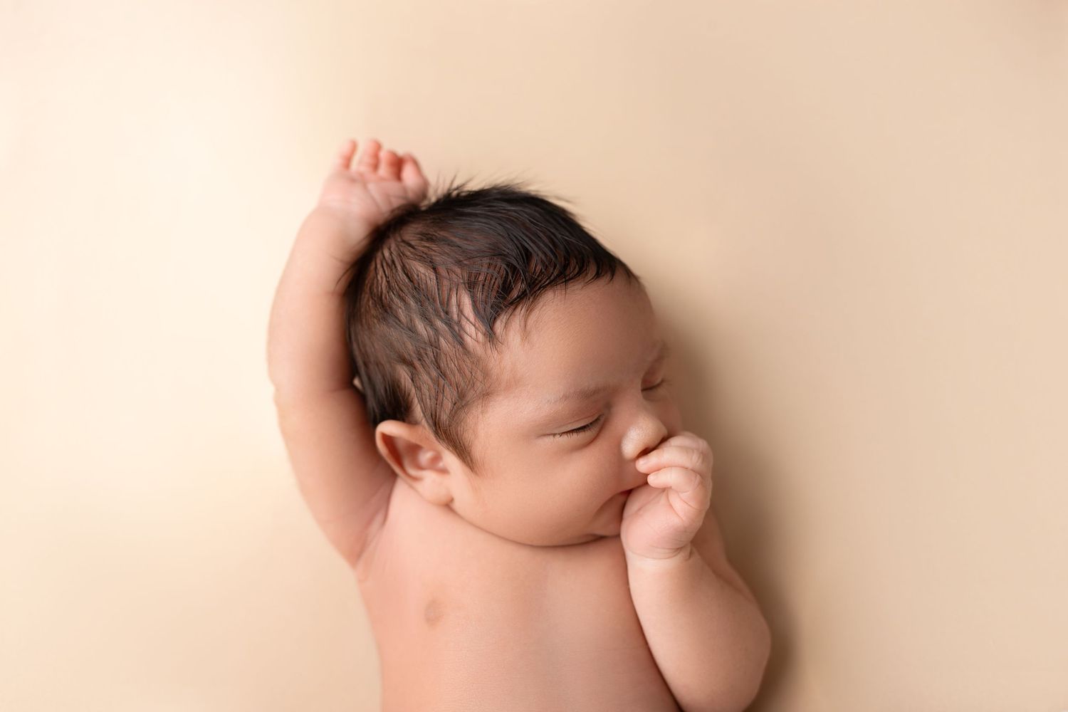 A peaceful sleeping newborn baby lying on a beige backdrop with one hand raised above their head.