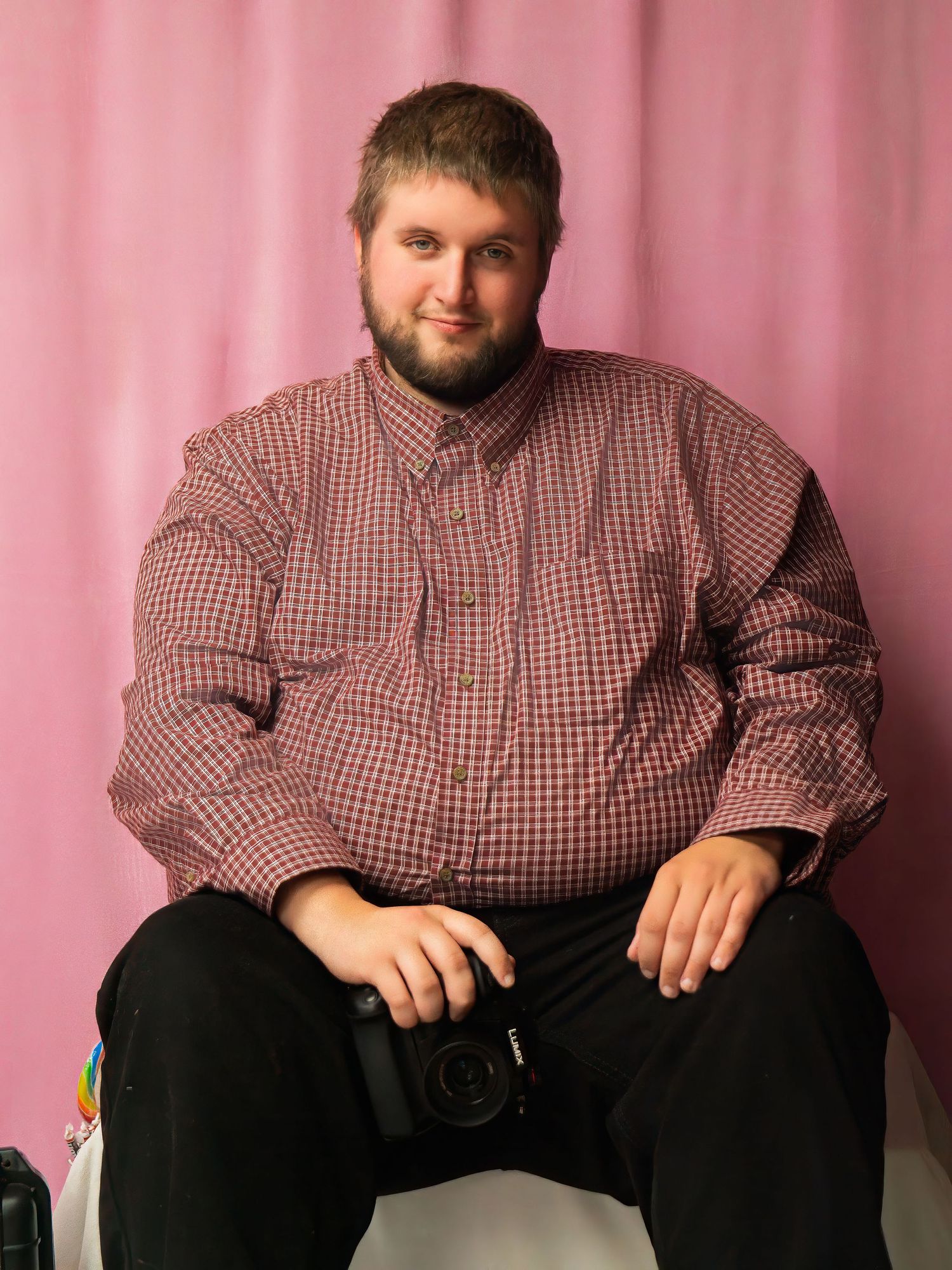 A photographer in a red checkered button-down shirt sits against a pink backdrop holding a camera.