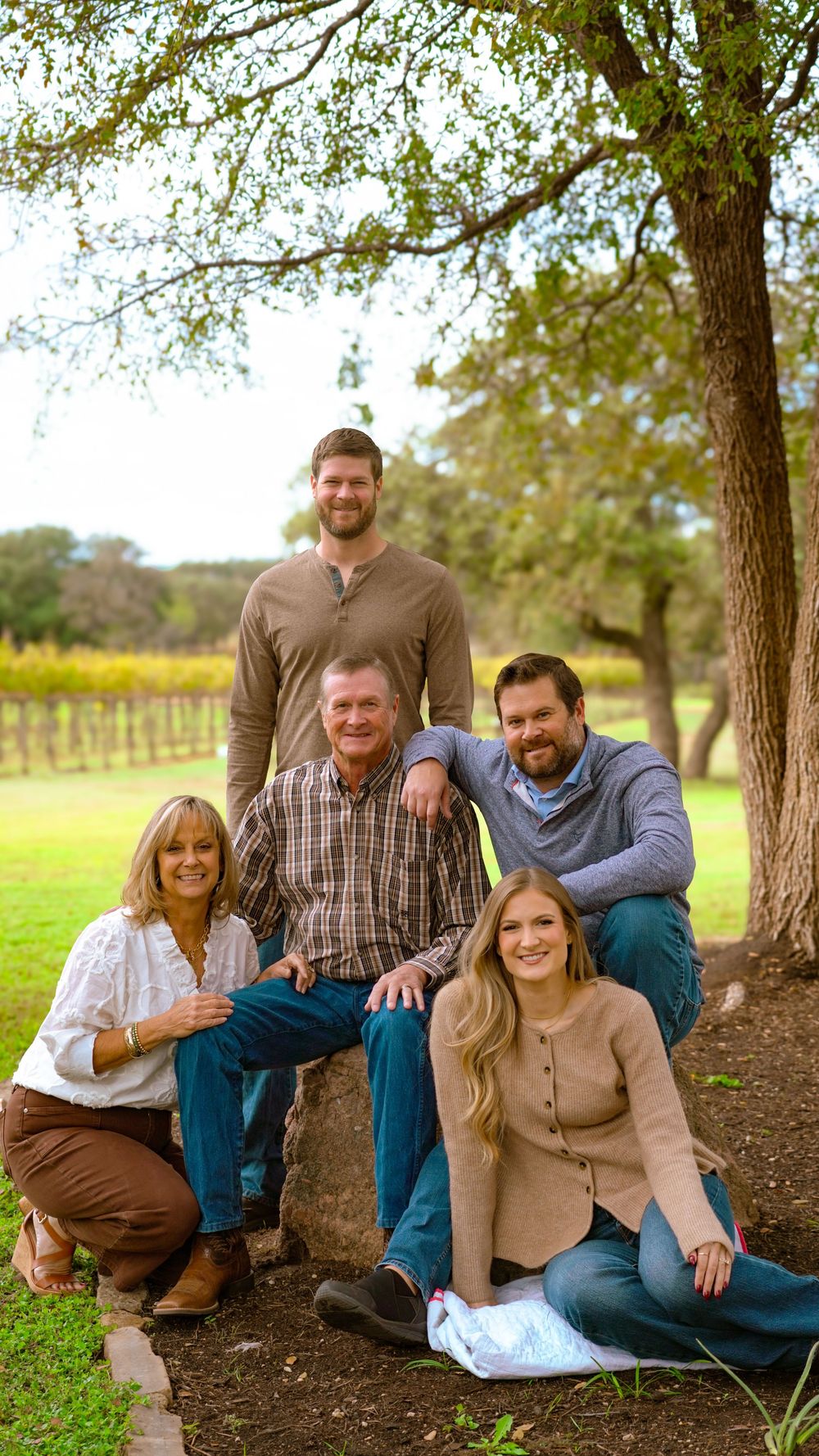 Family of five smiling together outdoors near a tree with vineyard in background on a sunny day.