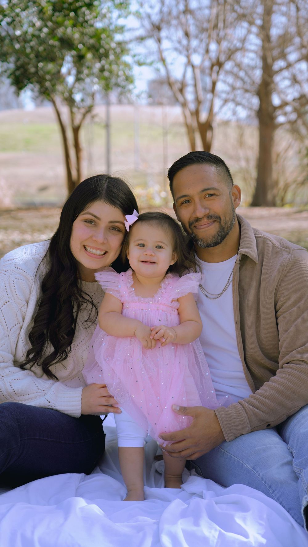 Happy family portrait outdoors, parents smiling with toddler girl in pink tutu dress seated between them.