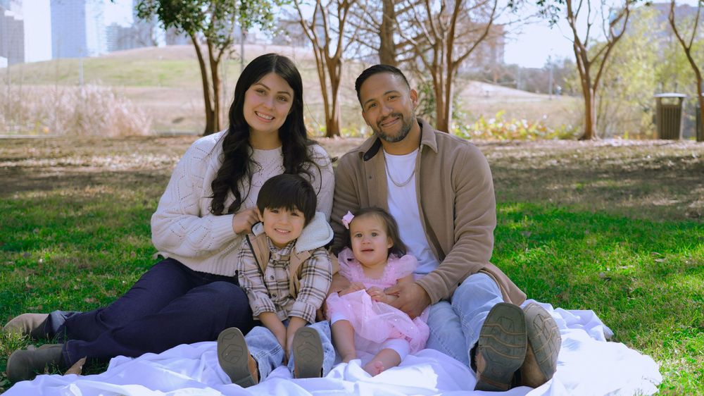 Happy family of four smiling together on a white blanket in a sunny park setting.