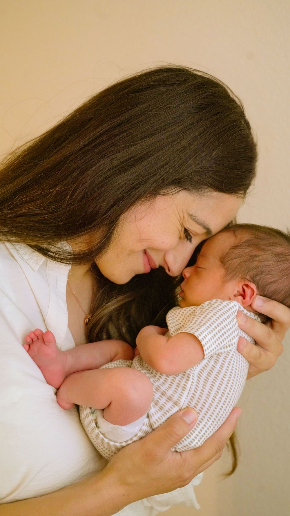 A tender moment of parent in white cradling a newborn baby wearing a checkered onesie against a neutral backdrop.
