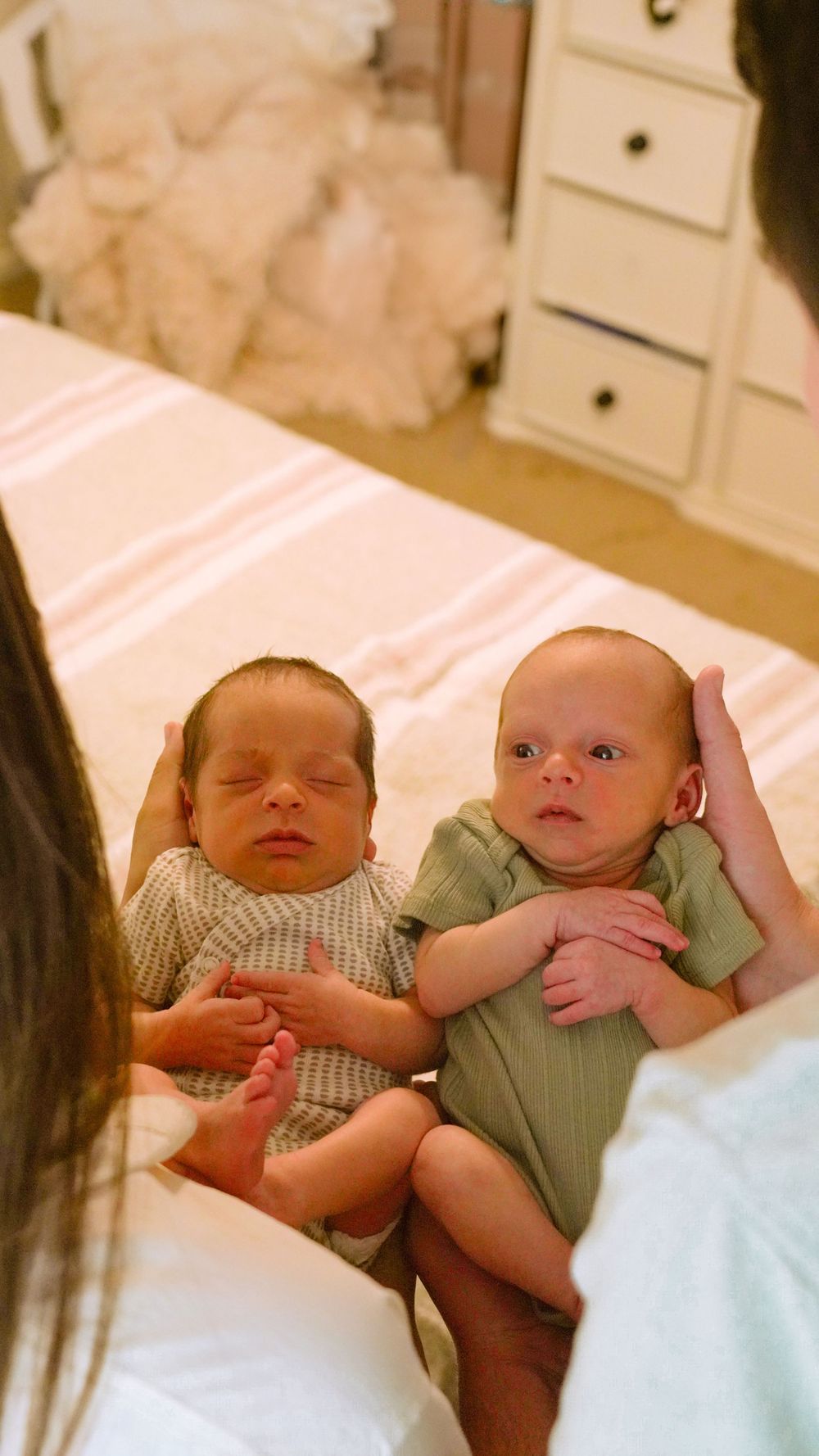 Two young babies in light colored onesies lying together on white bedding.