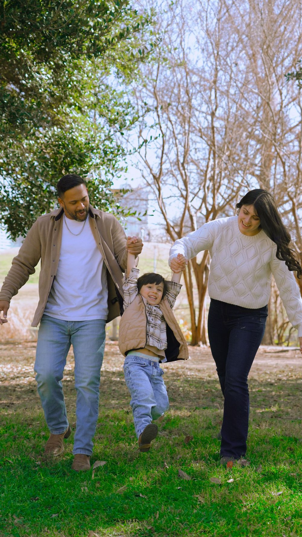 A joyful family of three walks outdoors, parents swinging their young child between them in a sunny park.