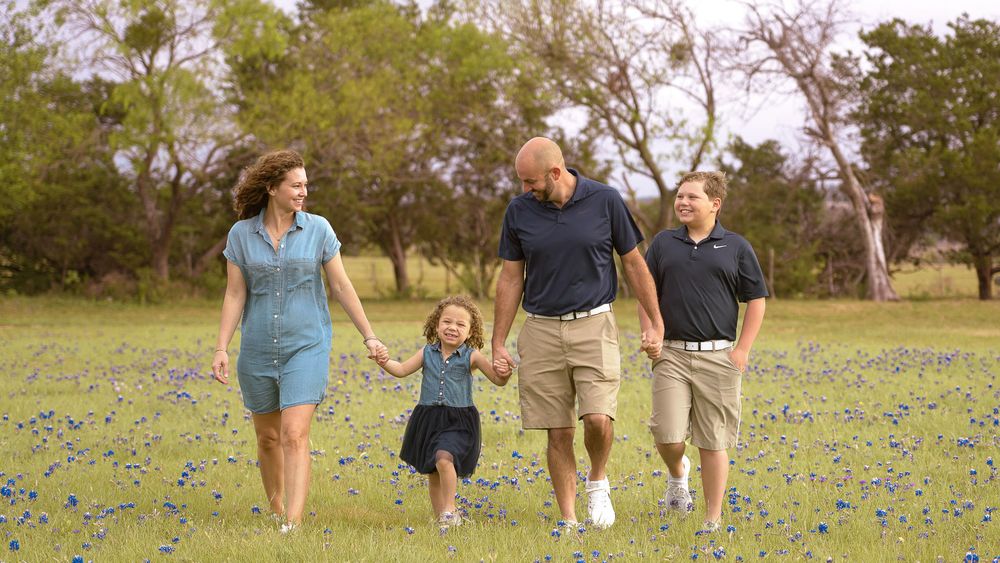 Family walking in a field of bluebonnets in Texas, smiling and holding hands on a sunny day.