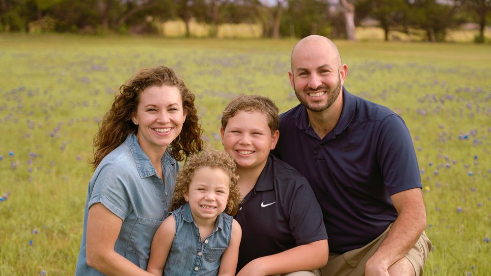 Family of four smiling together in a Texas bluebonnet field during a professional outdoor portrait session.