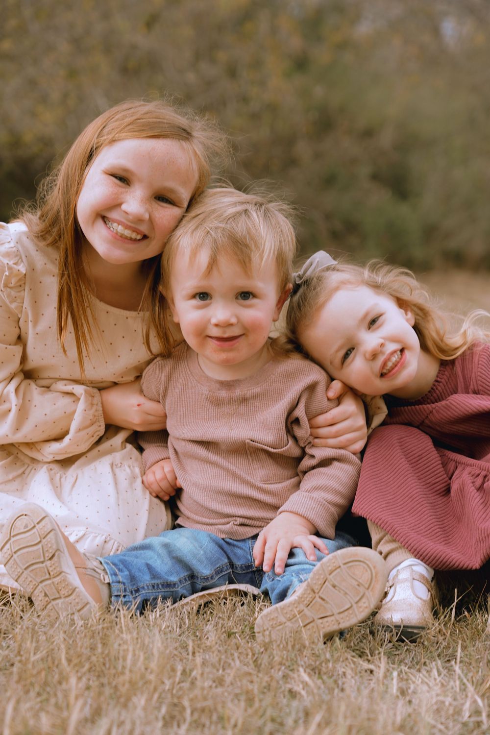 Three children sitting together in a field wearing cozy sweaters and smiling at the camera.