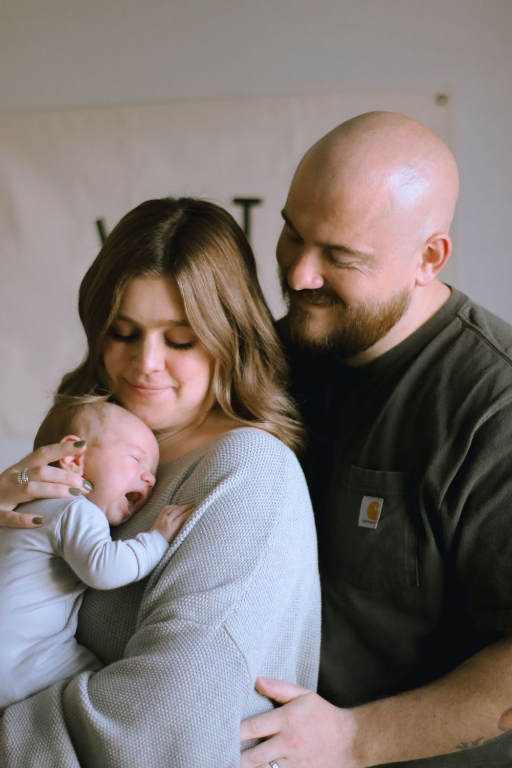 A young couple in casual attire holds their sleeping newborn baby in a tender family moment at home.