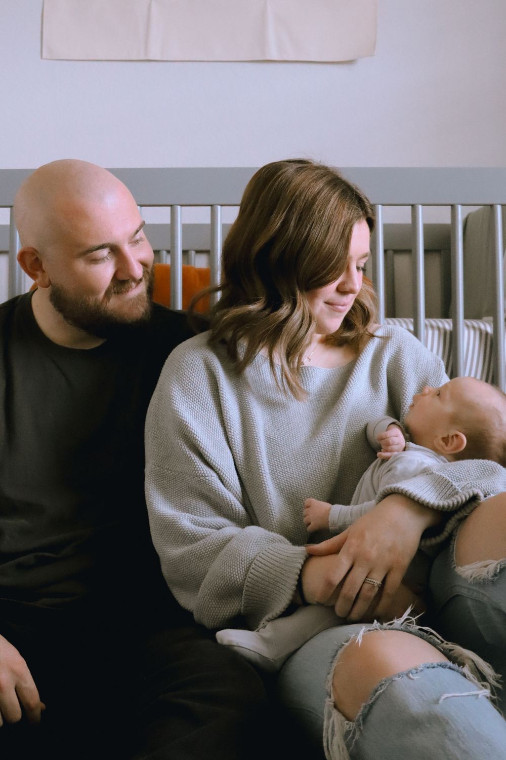A couple sits together on a couch with a baby, all wearing cozy sweaters in a tender family moment.