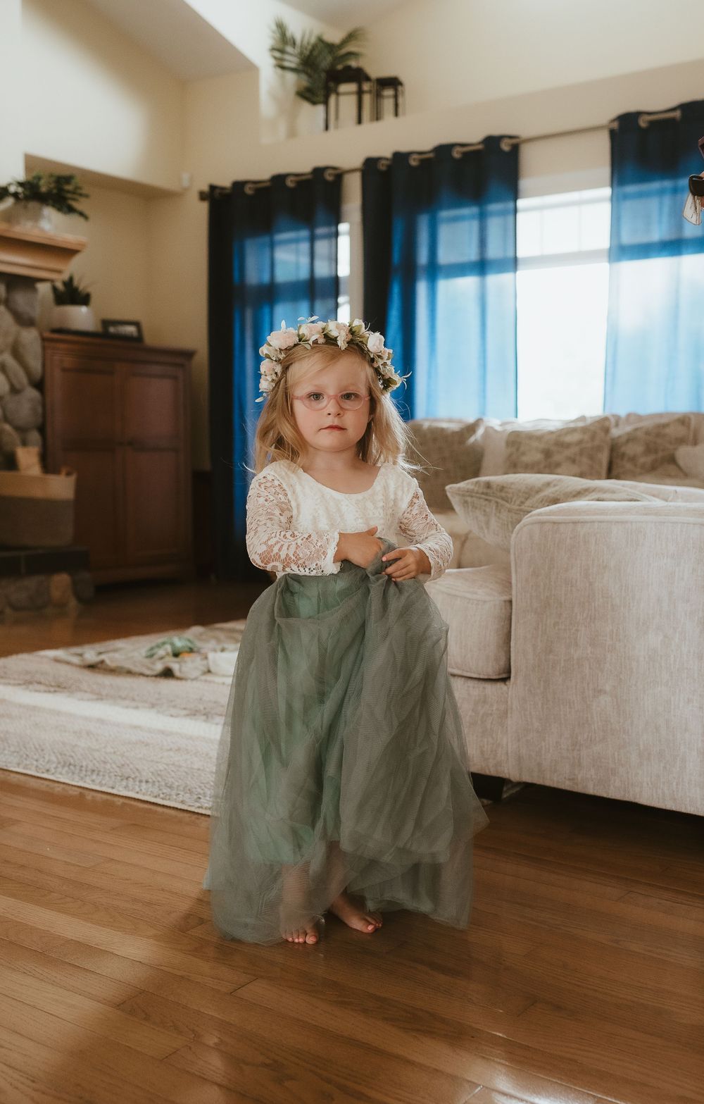 A small child wearing a floral crown, white lace top and sage green tulle skirt stands in a living room.