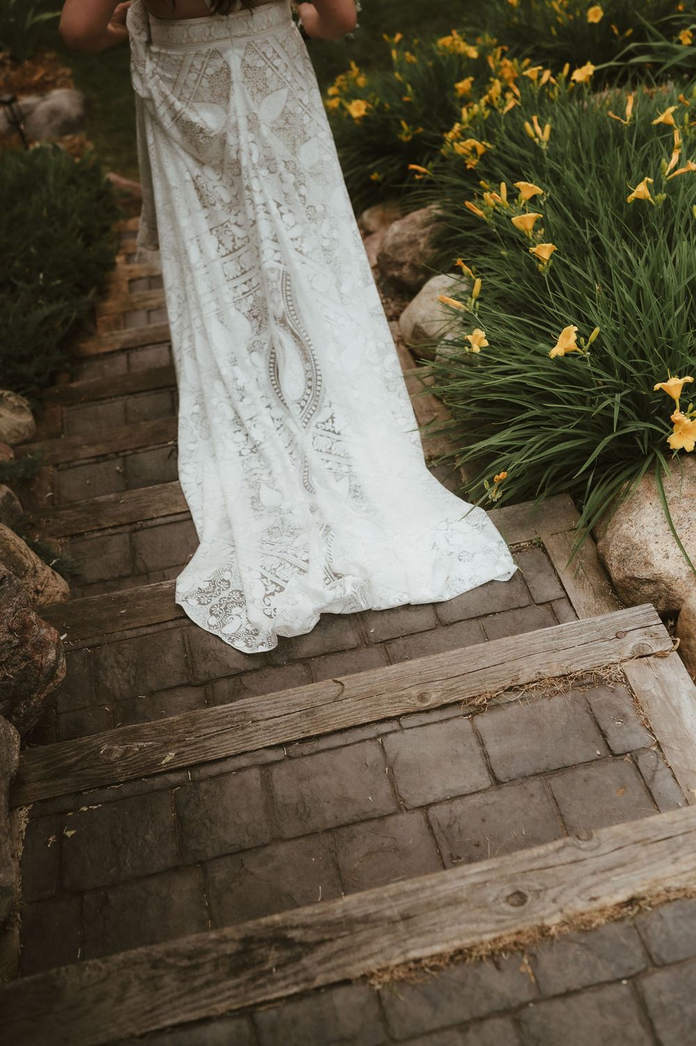 White lace wedding dress train flowing down stone steps near yellow daylilies.