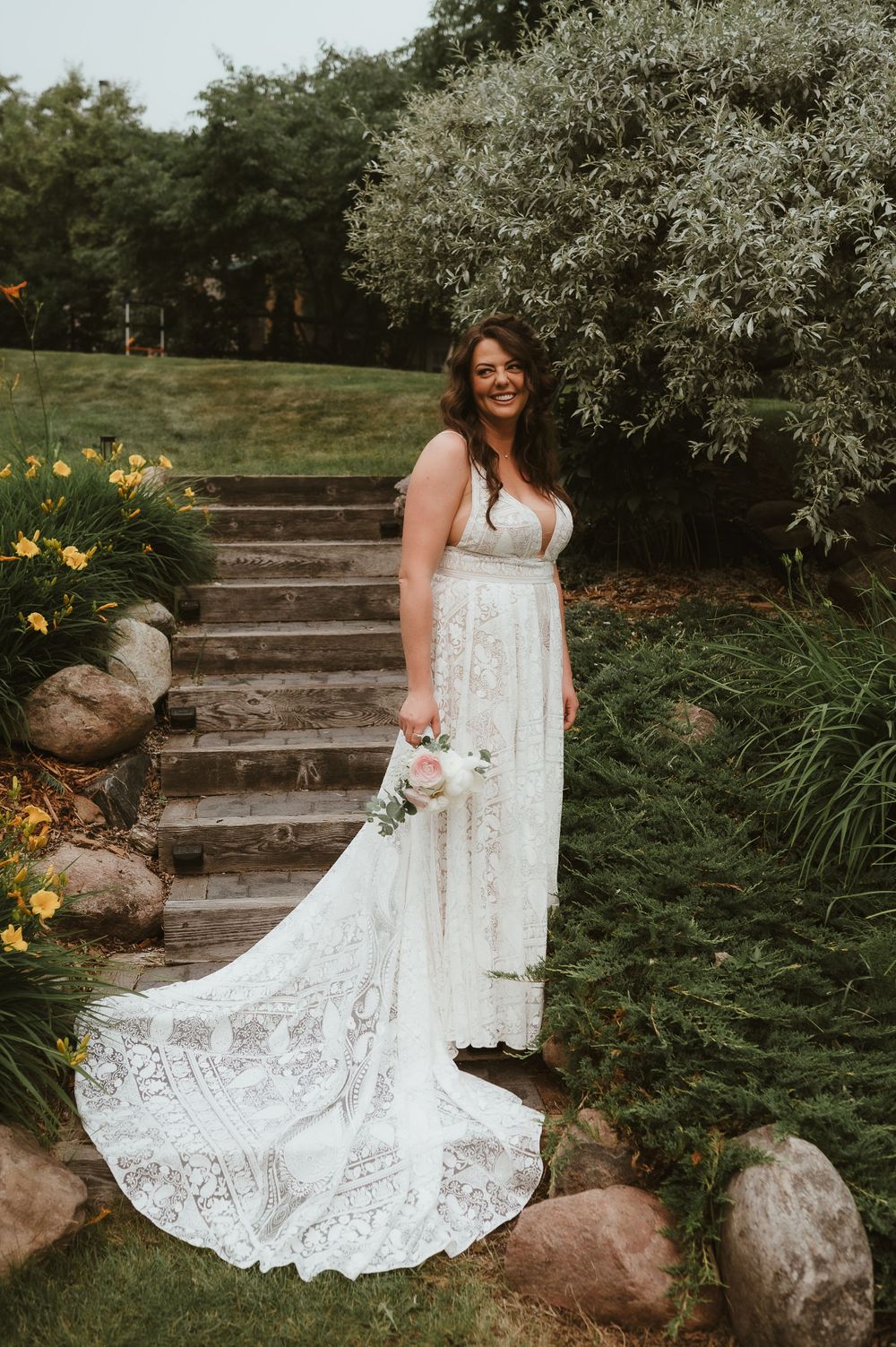 A bride in a lace wedding gown stands on stone steps surrounded by yellow flowers and greenery.