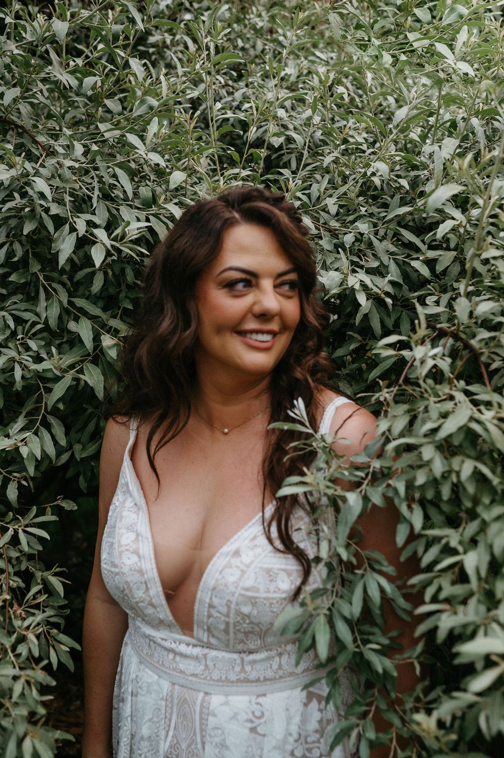 A woman in a white lace wedding dress poses among green foliage.