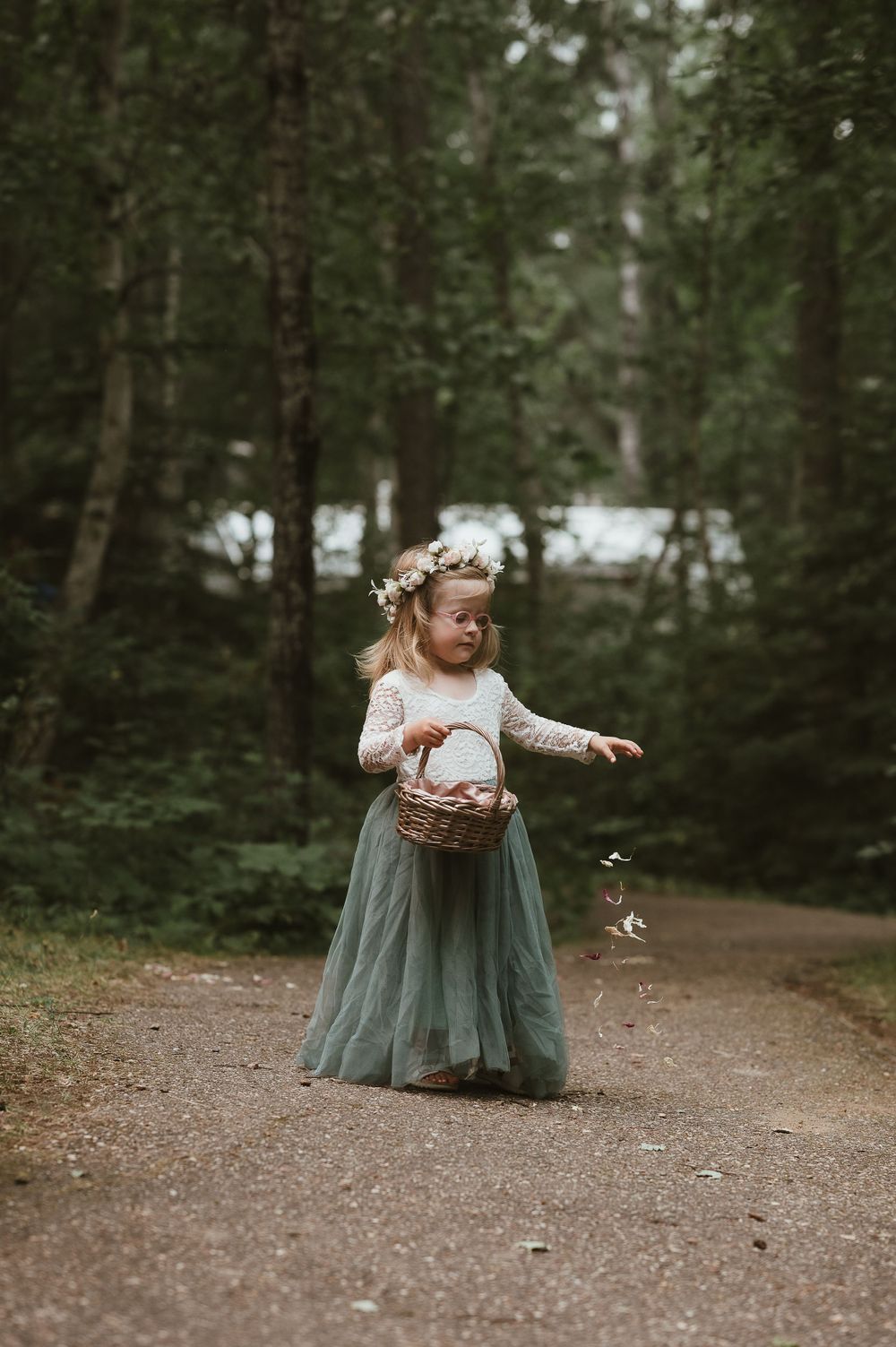 Young flower girl in sage green tulle dress carrying basket on forest path.