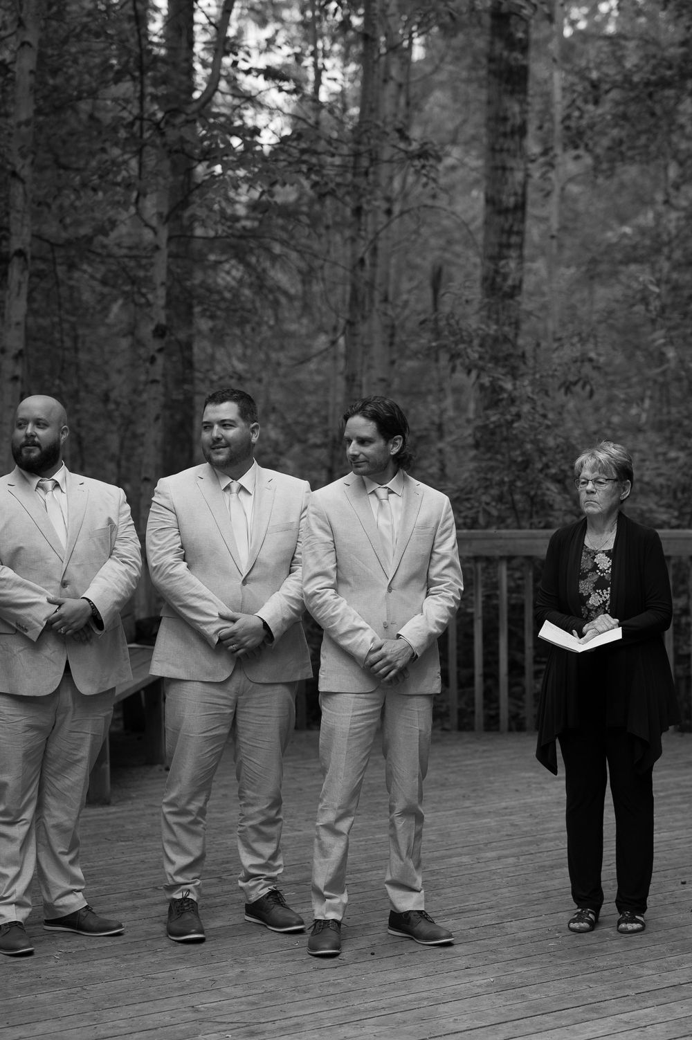 Groomsmen in light beige suits stand alongside an officiant on a wooden deck surrounded by forest.