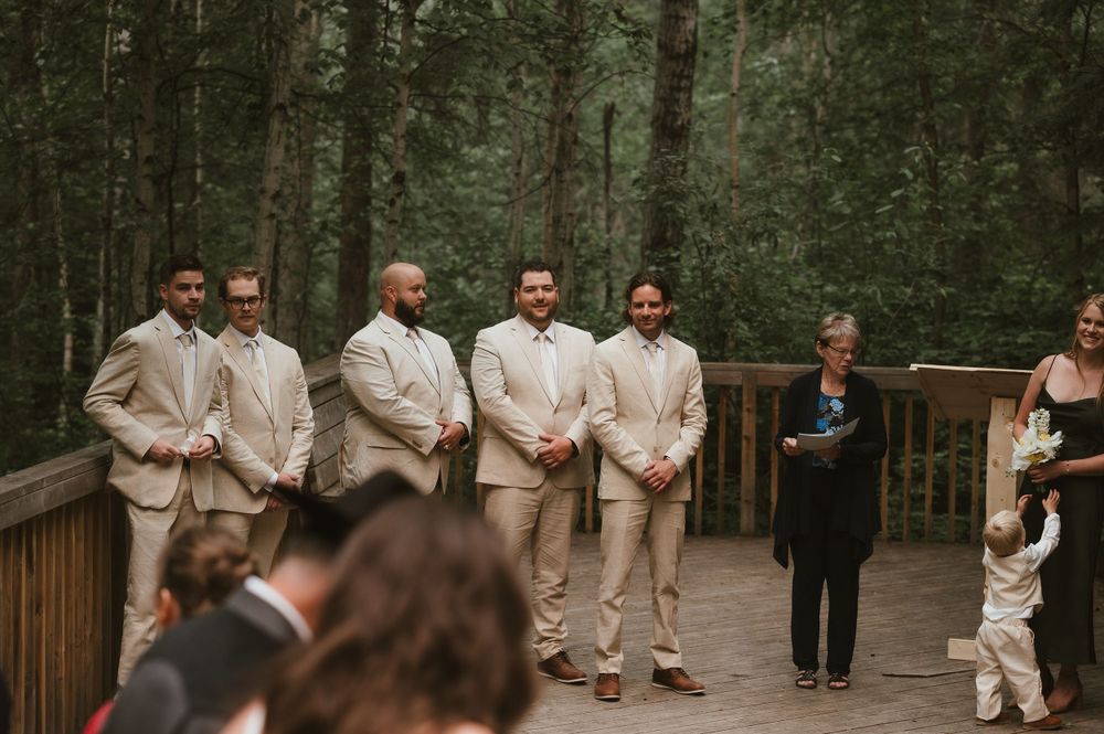 Wedding party in beige and dark attire gathered on a wooden deck in a forest setting.