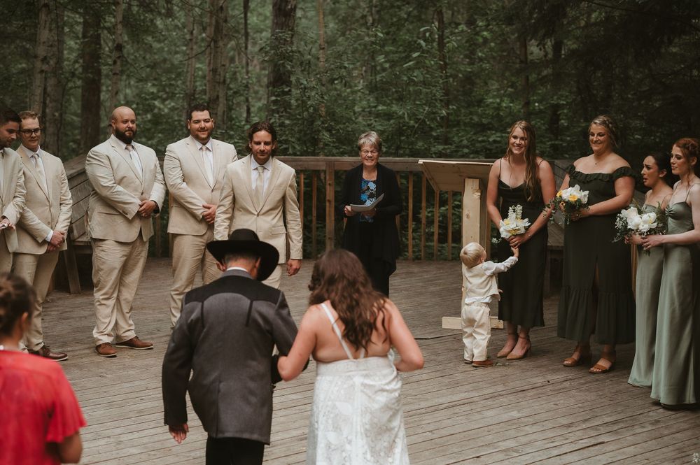 Forest wedding ceremony in progress with wedding party in beige suits and dark dresses on wooden deck.