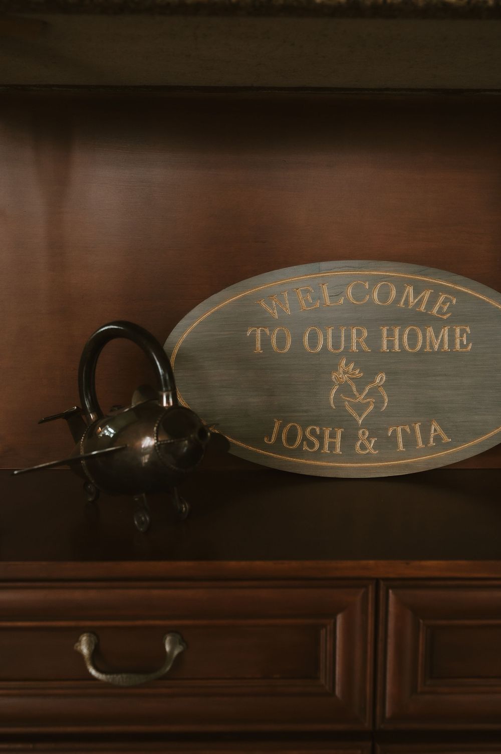 A wooden welcome sign and teapot sit on a dark wood shelf in warm lighting.