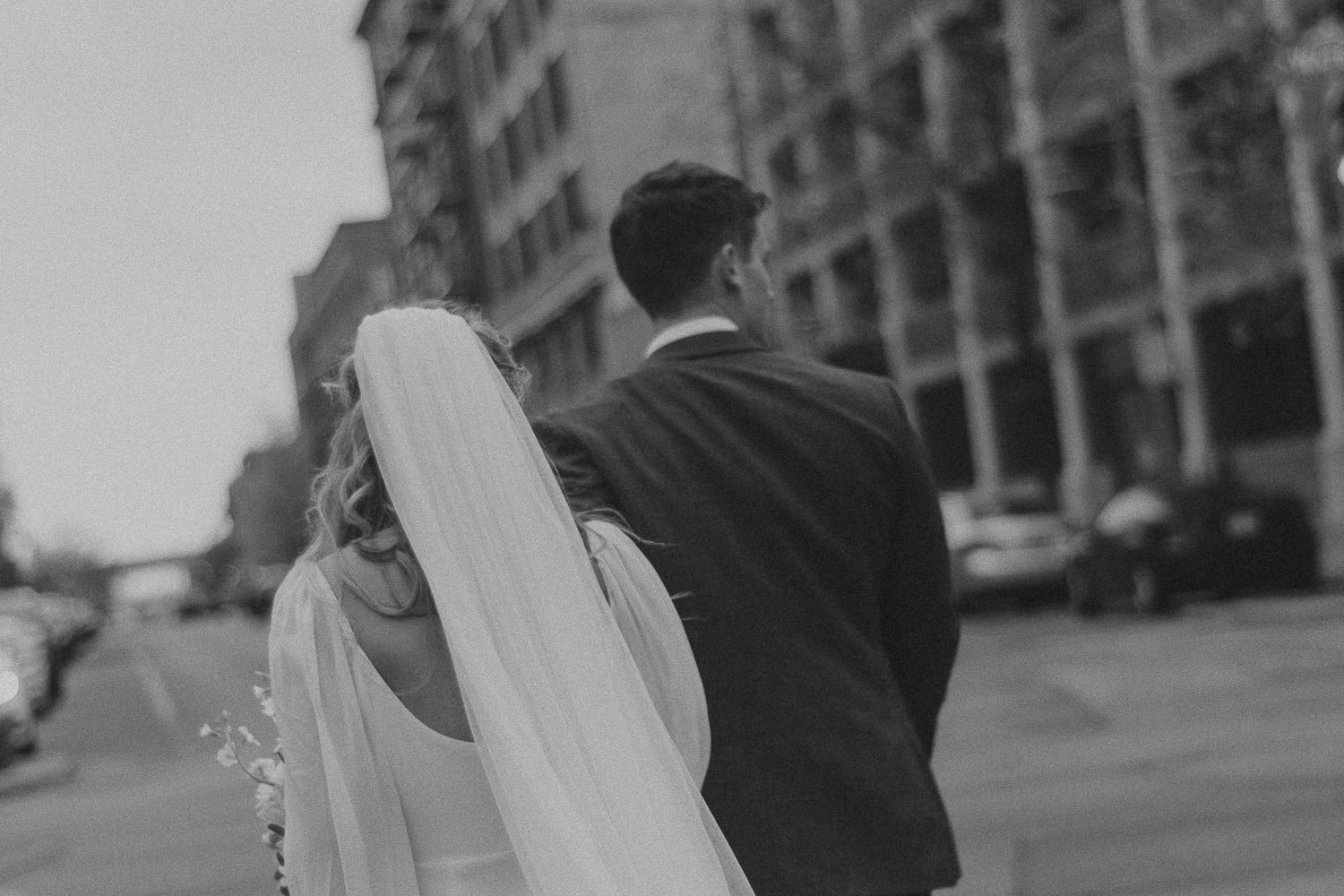 Black and white photo of a bride's veil and groom's back as they walk down a city street with tall buildings.