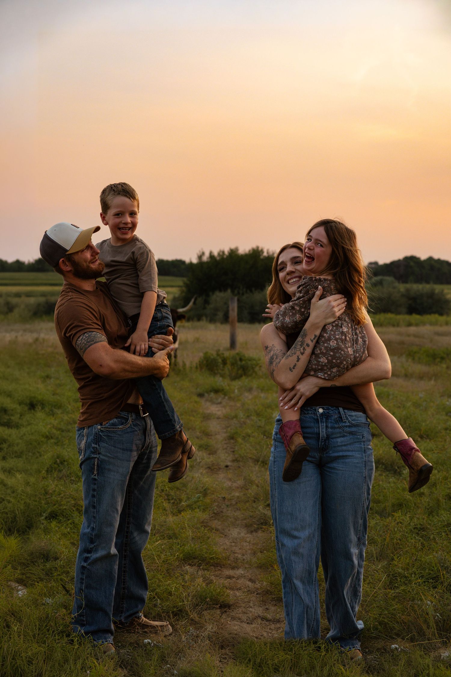 Happy family of four with two young children laughing together in green field at golden sunset.