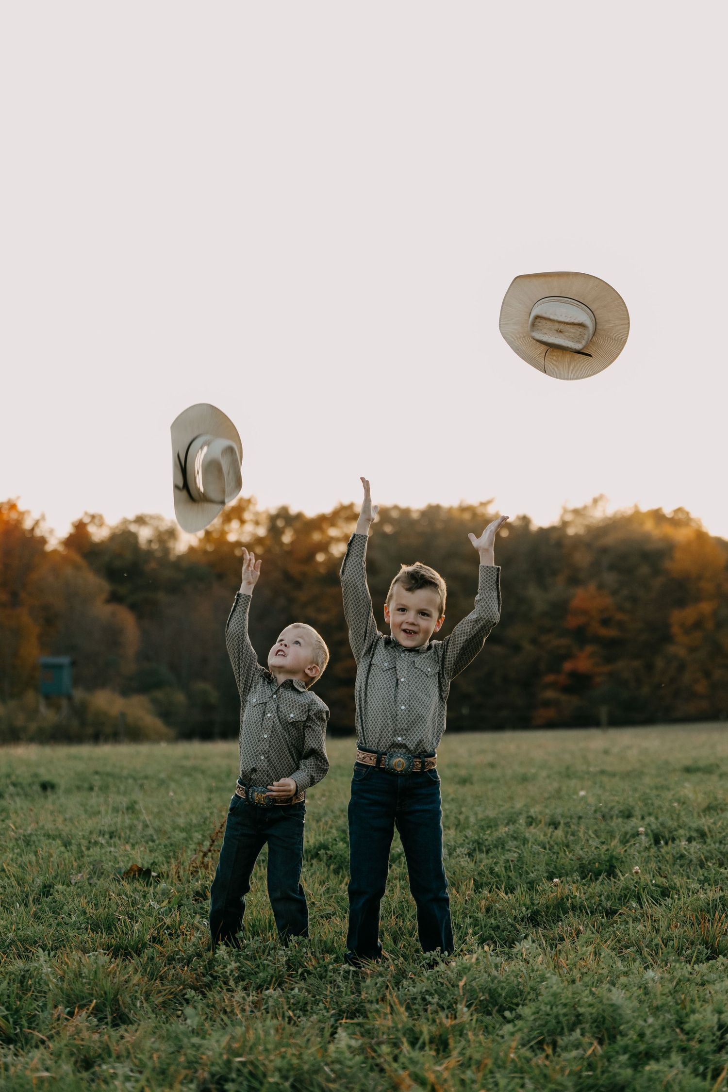 Two young children toss cowboy hats in the air while playing in a grassy field during golden hour.