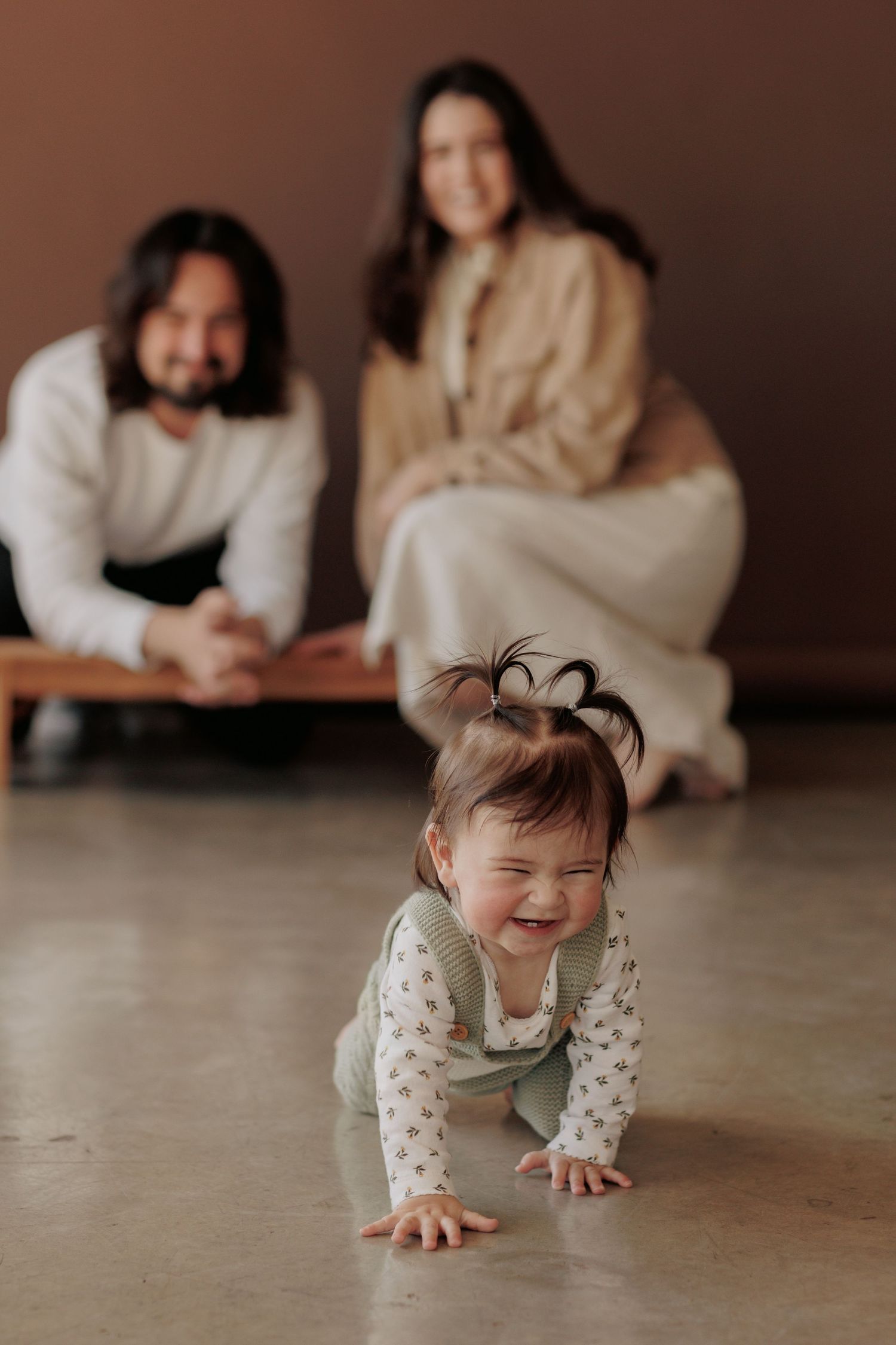 Joyful baby crawls on floor while parents watch from behind in cozy indoor setting.