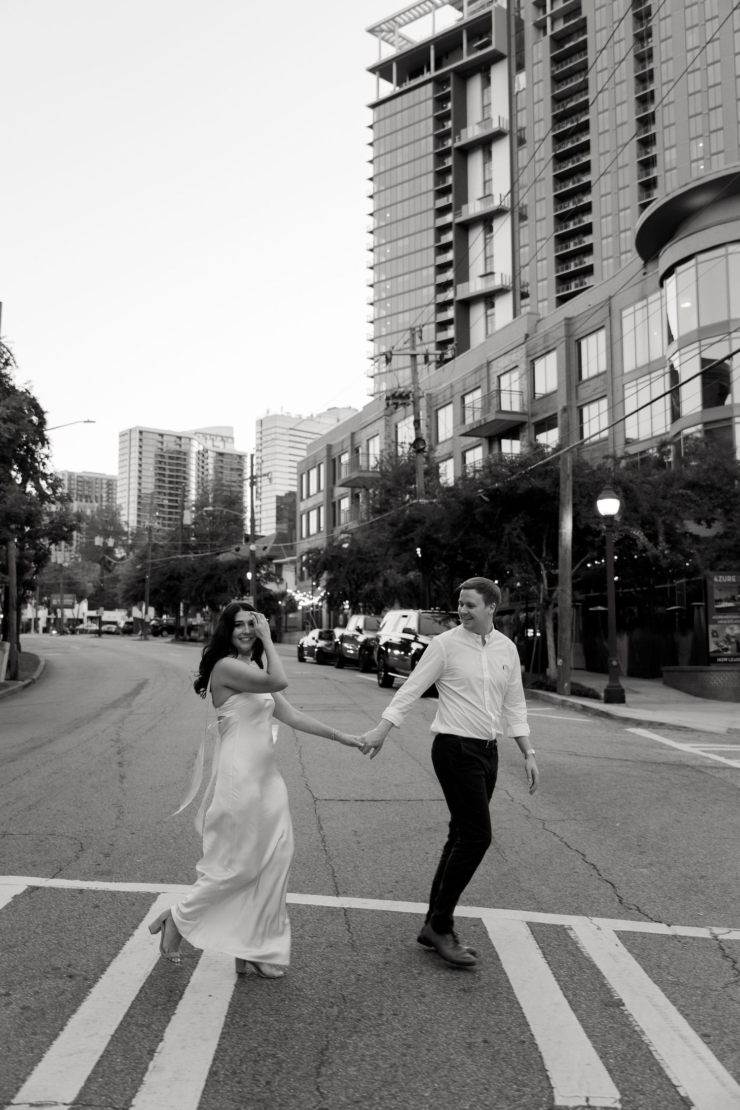 A couple in formal attire joyfully crosses a downtown city street with high-rise buildings in the background.