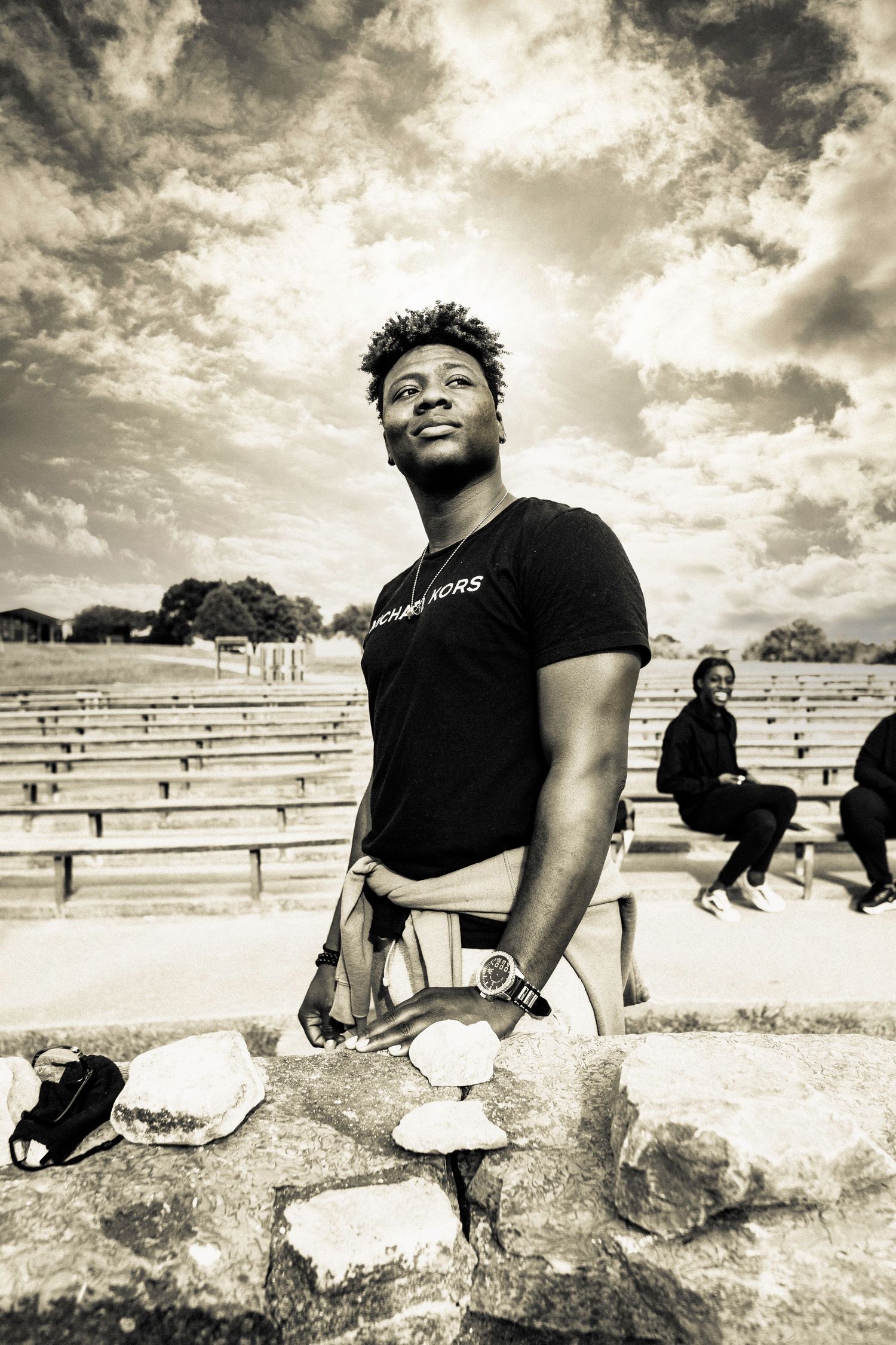 Black and white photo of someone standing outdoors in a t-shirt in front of stadium bleachers under dramatic clouds.