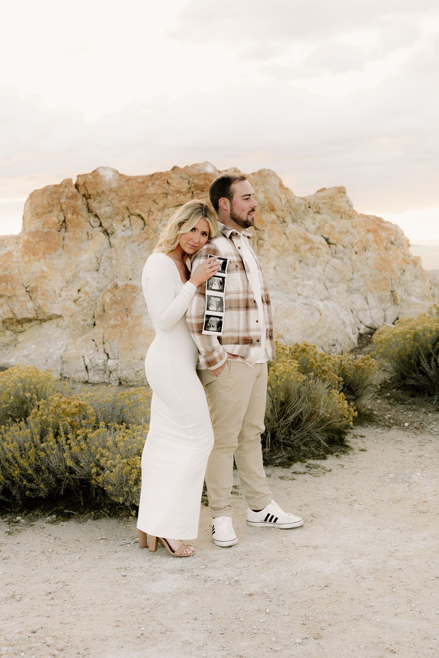 A couple in formal attire embraces while standing in front of a rocky desert landscape at sunset.