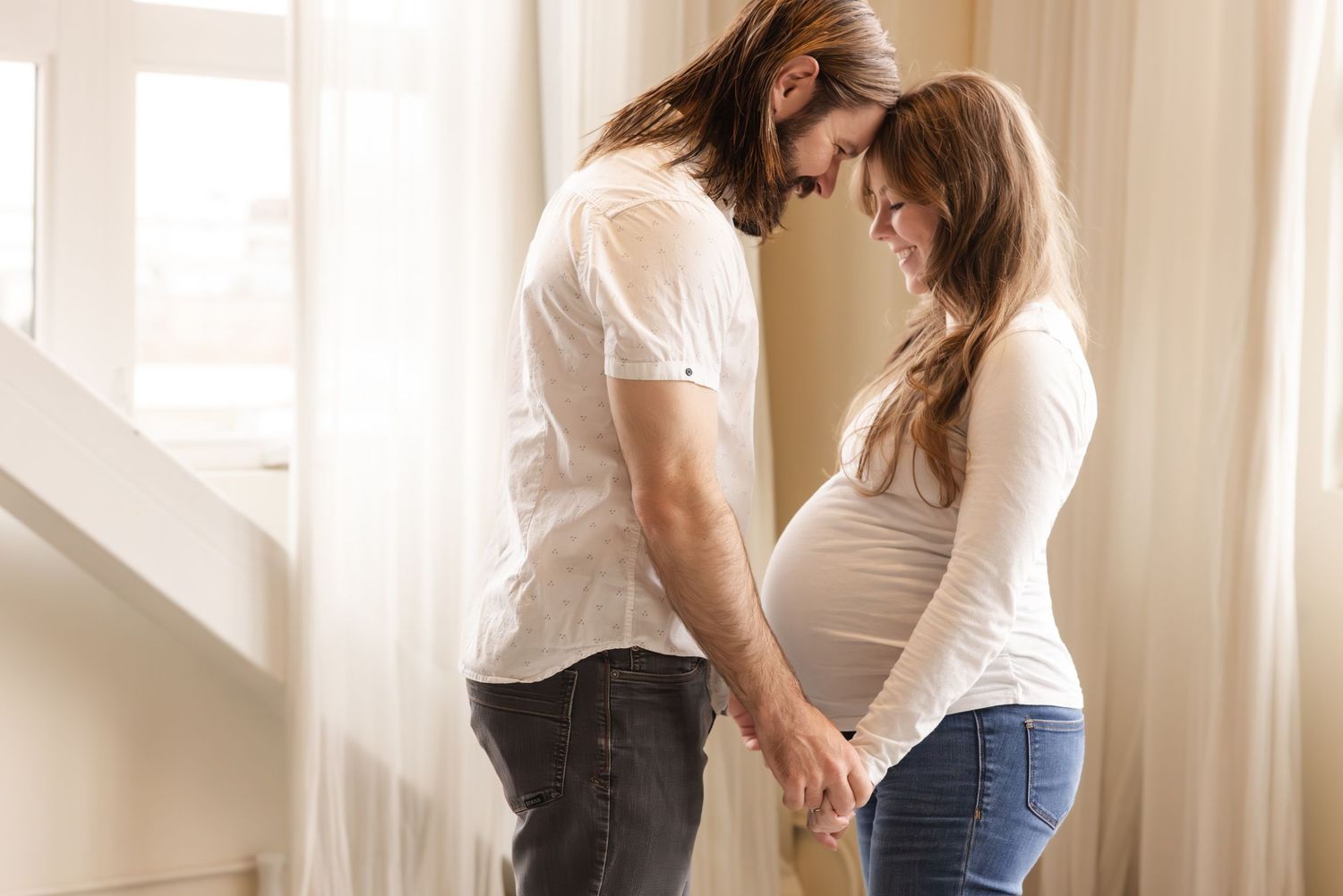 A couple stands together by a window, with an expectant mother showing her pregnant belly in a tender moment.