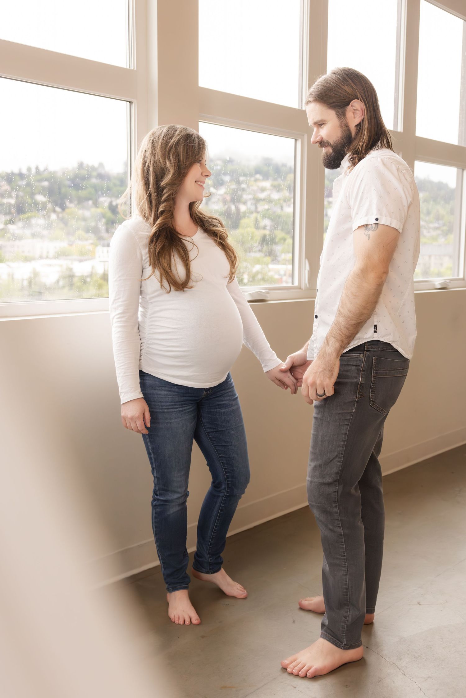 A couple stands barefoot holding hands near tall windows, with the person on the left wearing a white maternity top.