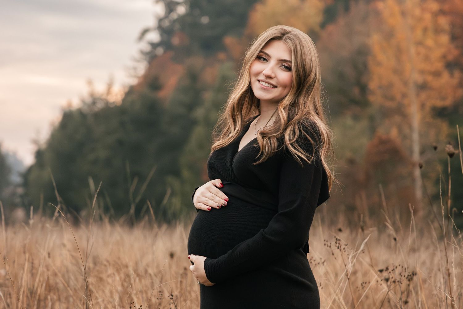 Expectant mother in black sweater poses in autumn field with tall grass and fall foliage.