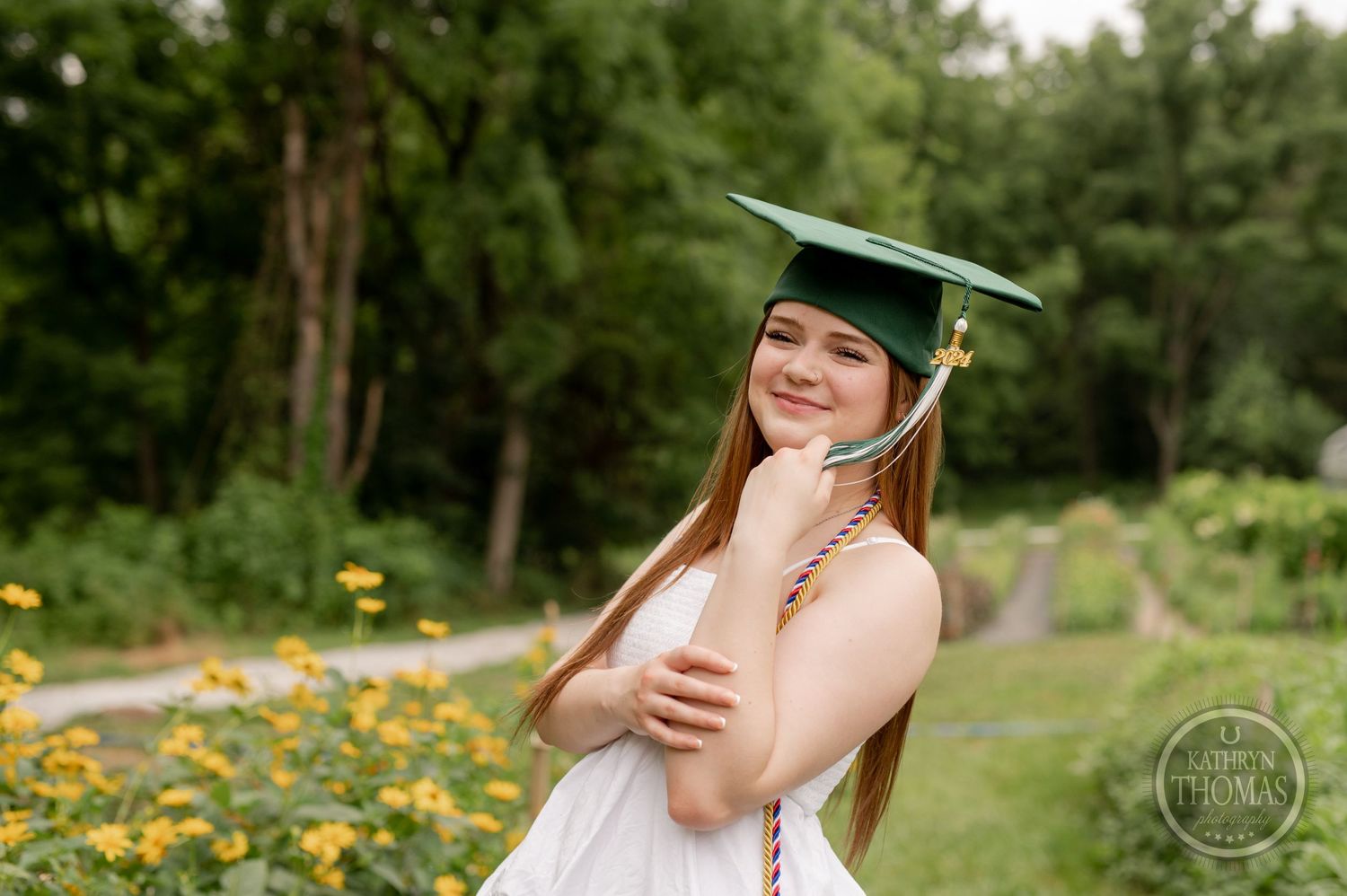 Graduate in green cap and white dress poses joyfully beside yellow flowers in garden.