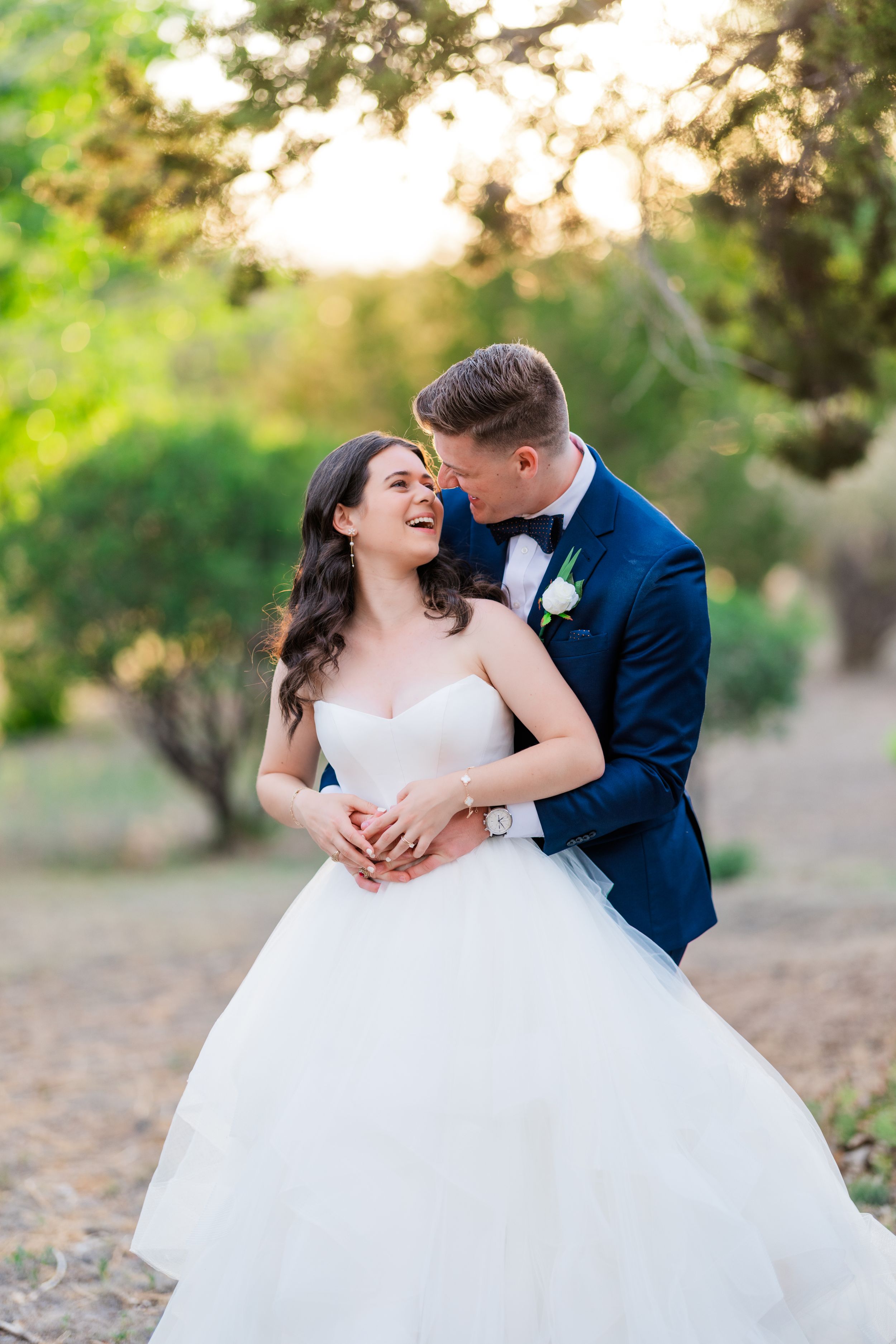Romantic wedding couple embraces outdoors at sunset surrounded by soft bokeh and greenery.