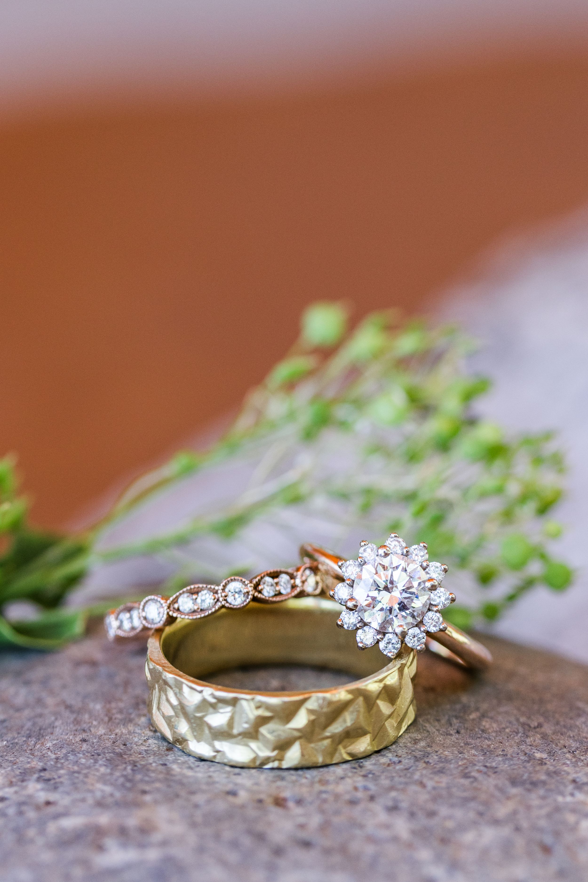 Two wedding rings on textured stone surface with delicate green foliage, featuring diamond clusters and gold band designs.