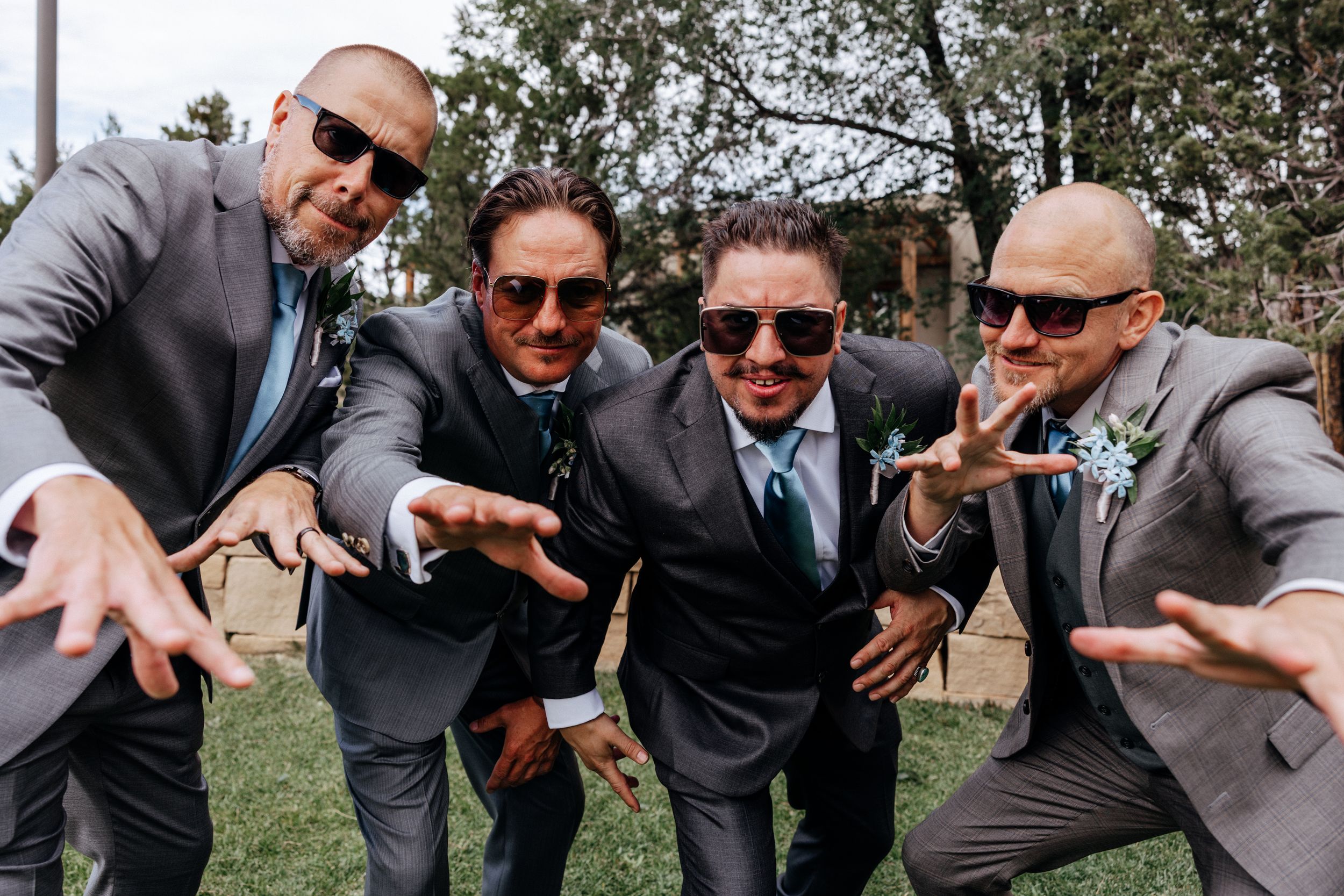 Groomsmen in matching suits and sunglasses strike playful poses on grass during outdoor wedding celebration.